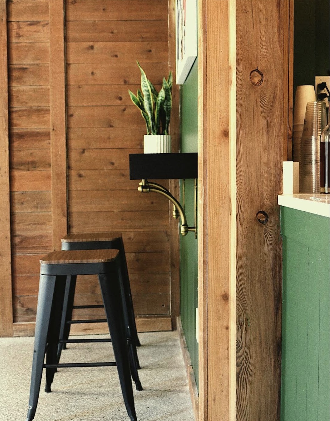 Interior view of a wooden wall with a small black shelf holding a potted plant, two black stools, and part of a green wall.