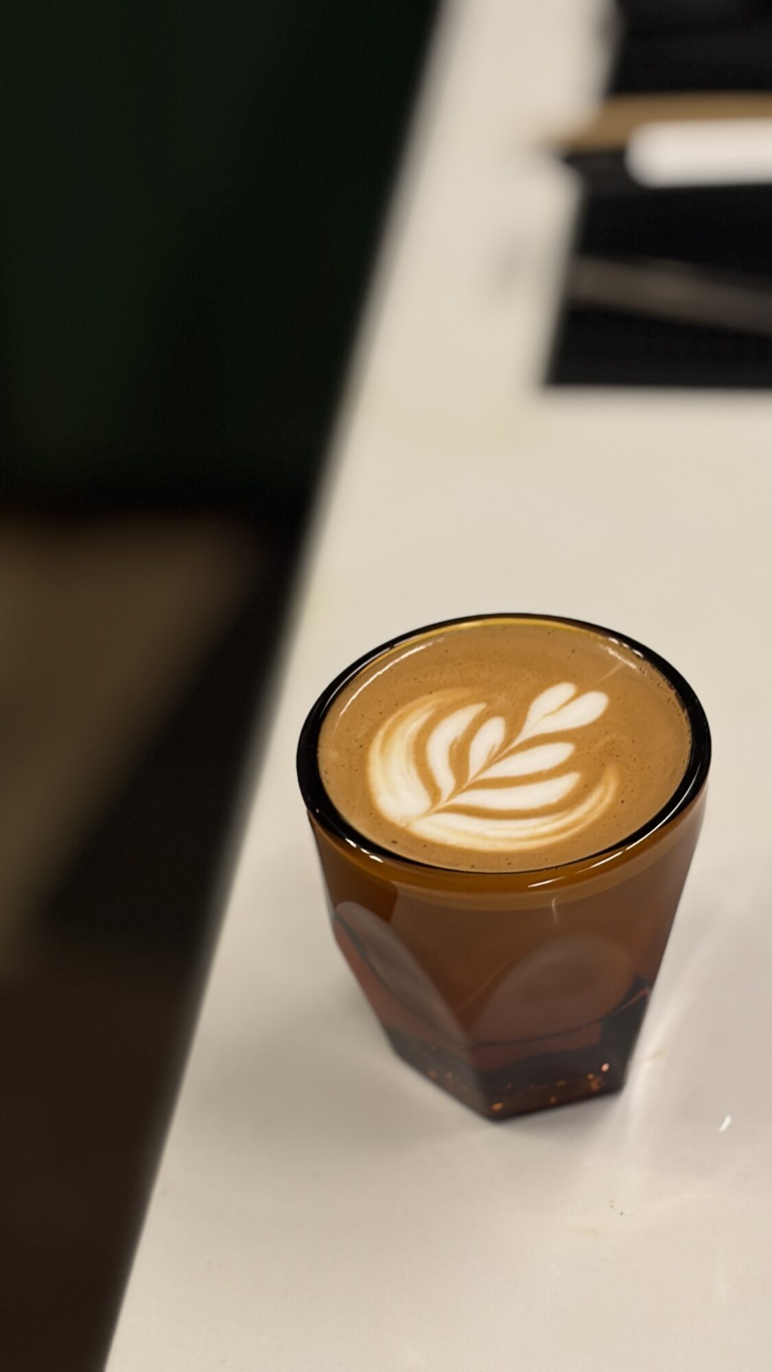 Glass of coffee with latte art on top, placed on a white surface.
