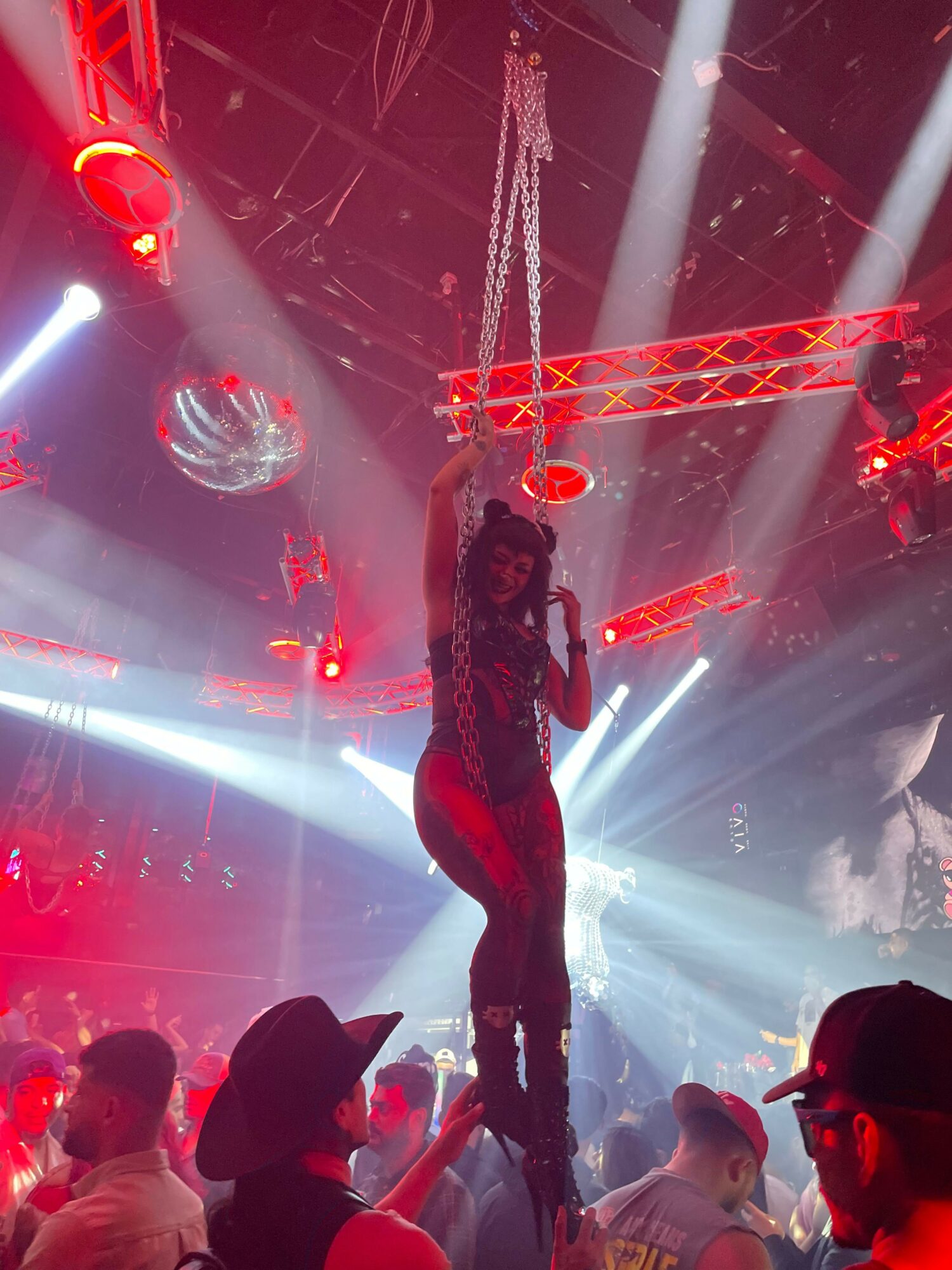 Performer hanging from a chain on stage with colorful lights and audience in foreground.