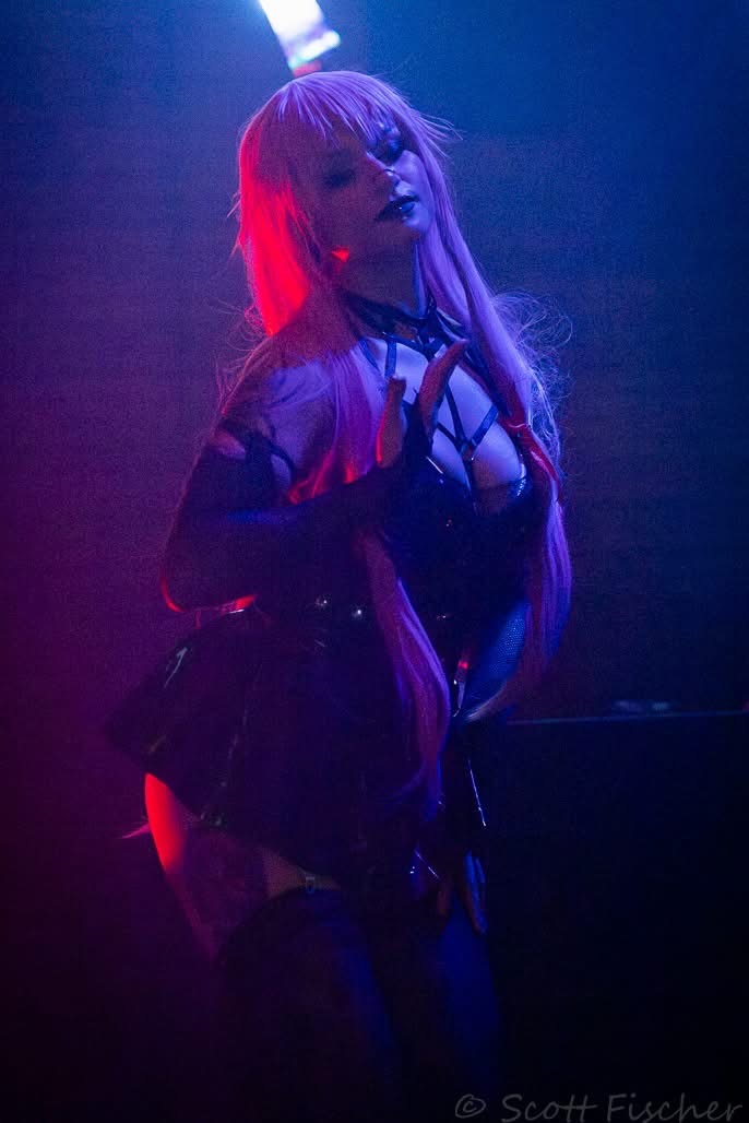 Person with long hair in a dark outfit, illuminated by purple and red lighting, standing in a dim environment.