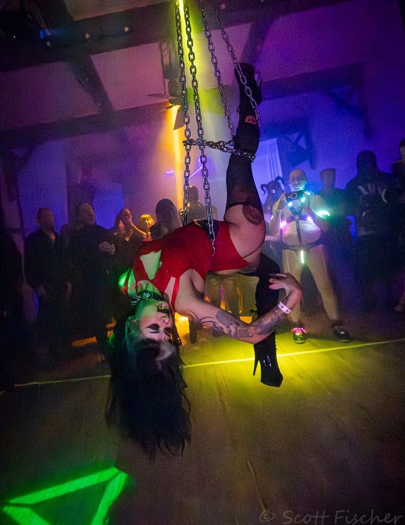 Performer hanging upside down from chains in a nightclub with colorful lighting and a crowd watching.