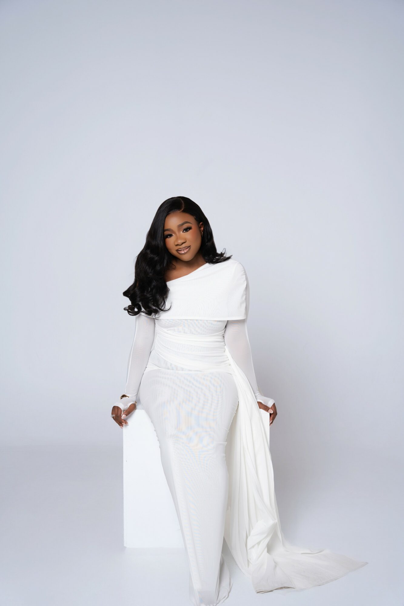 Woman in white dress sitting on a white block against a plain light gray background.