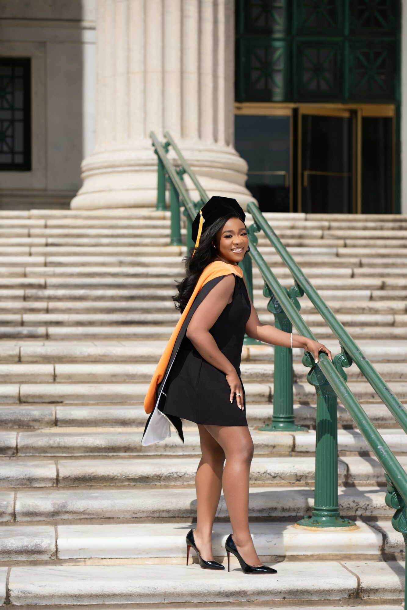 A woman in a black dress and high heels stands on steps in front of a building with large columns, smiling.