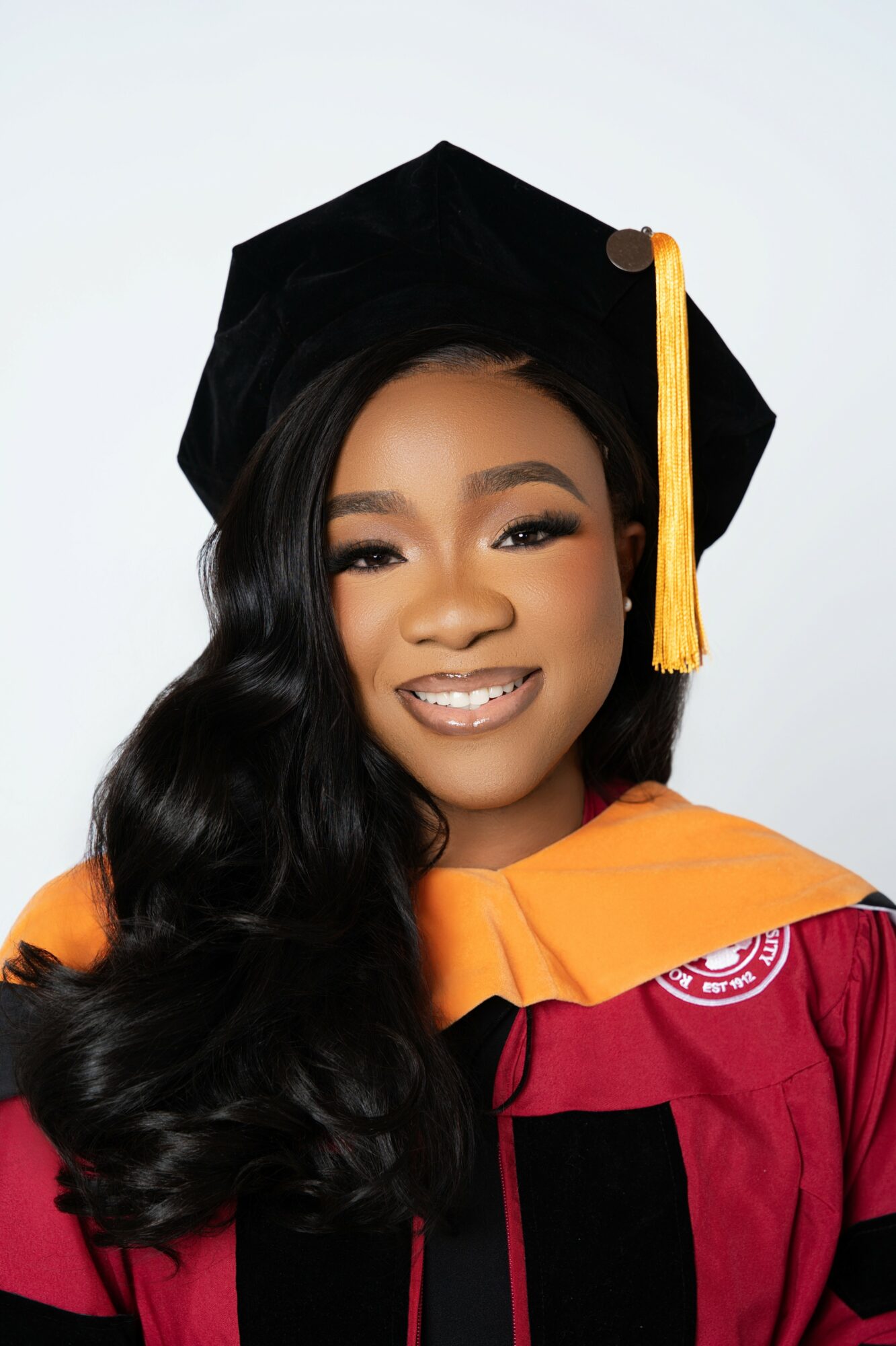 Young woman in graduation cap and gown smiling, with long wavy hair and makeup, against a plain background.