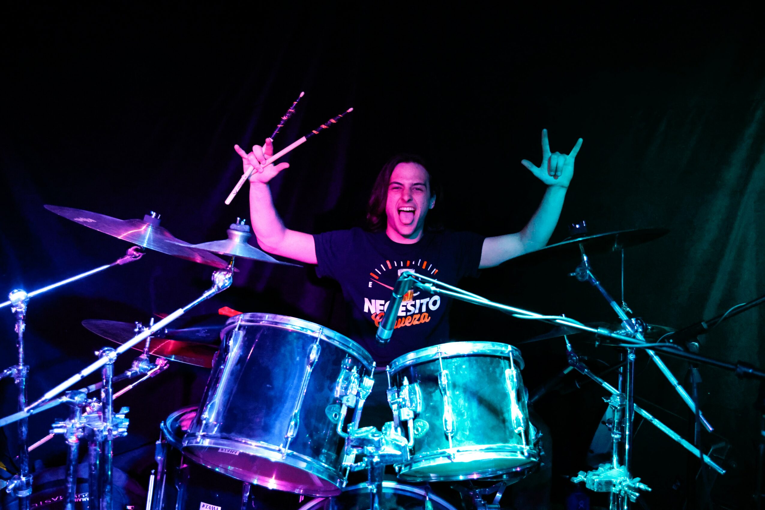 Person playing drums on stage, smiling, with hands raised, surrounded by drum set and stage lighting.