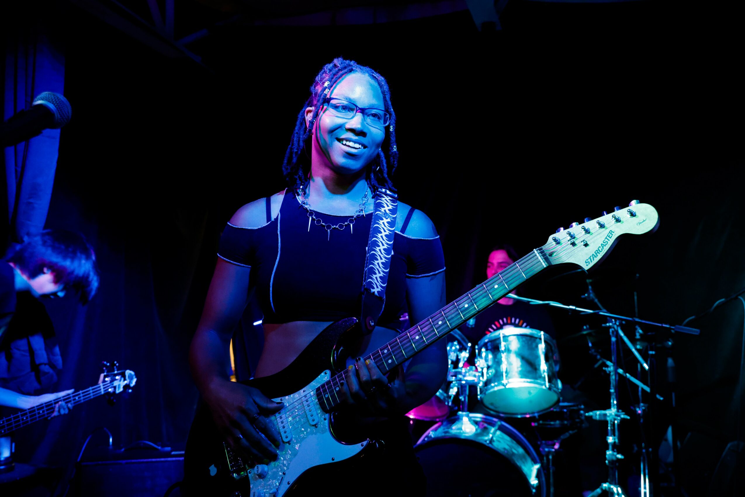 Person smiling while playing electric guitar on stage with drums in background, dark lighting with blue highlights.