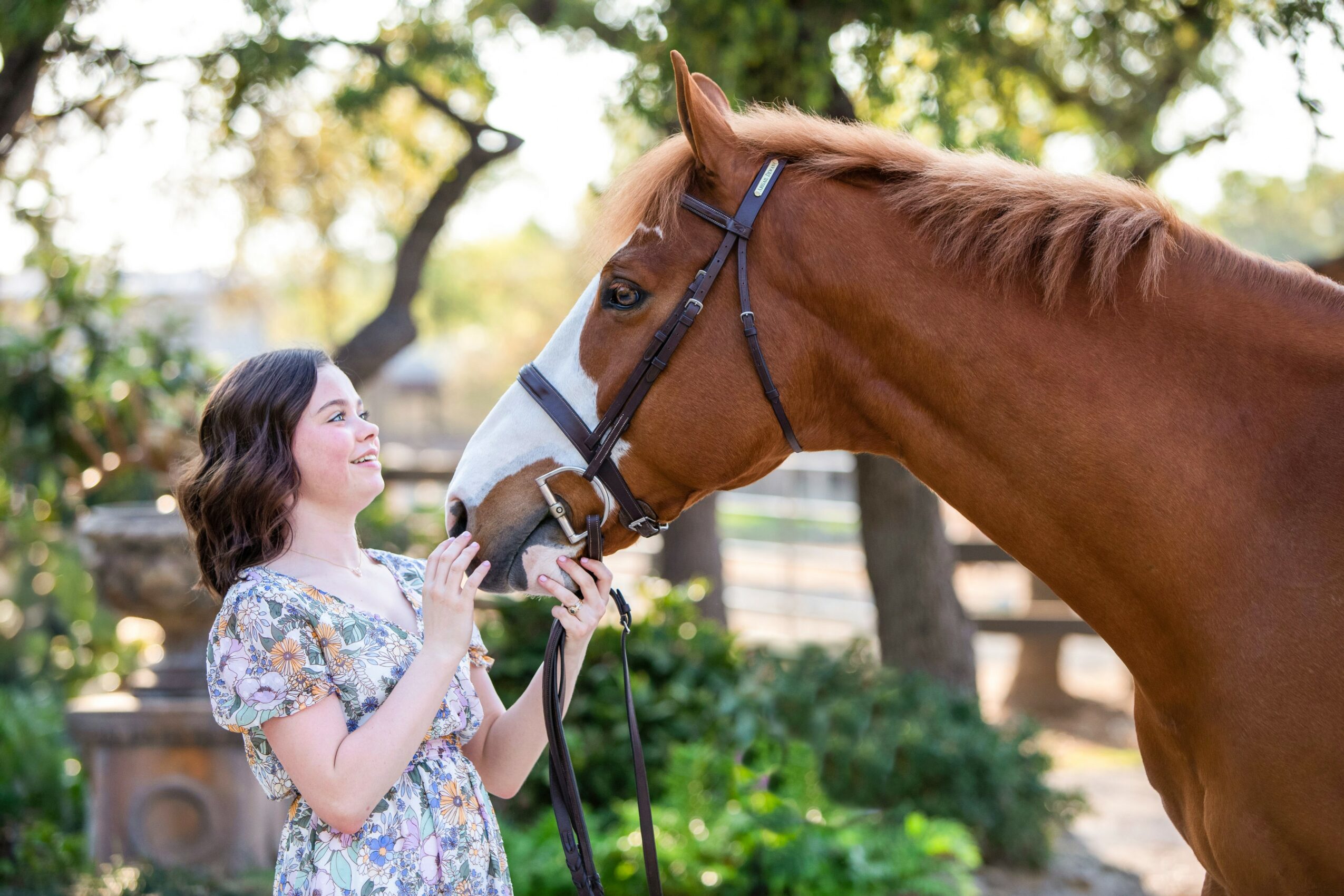 Girl in floral dress holding a horse's reins outdoors with trees and greenery in background.