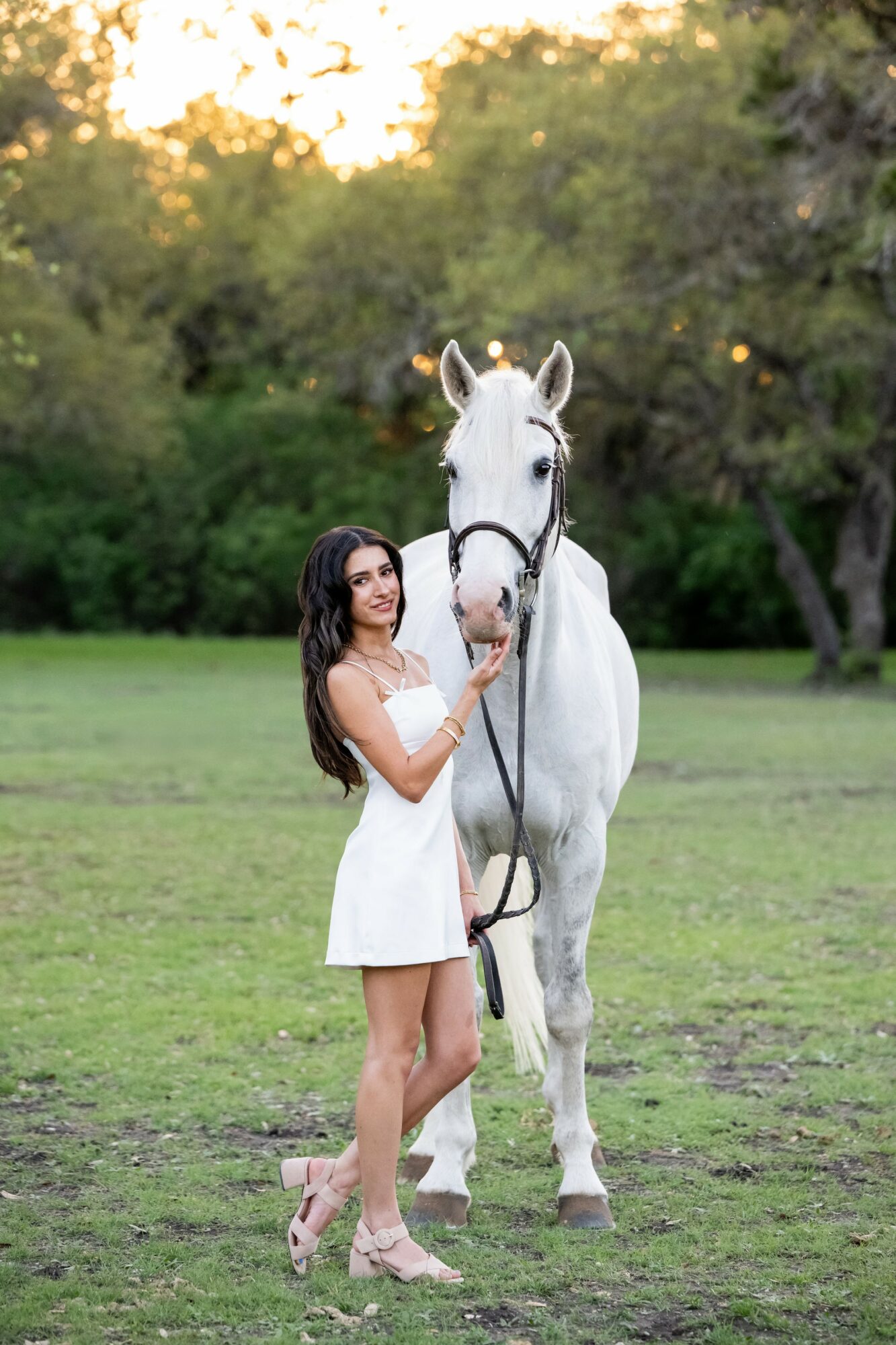 Woman in white dress standing next to a white horse in a grassy field with trees in the background.