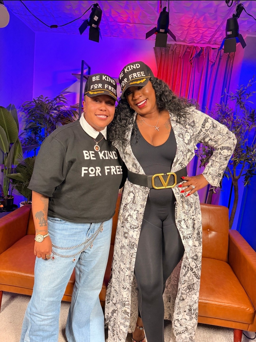 Two women standing together, smiling, in a room with purple and pink lighting, plants, and orange couches.