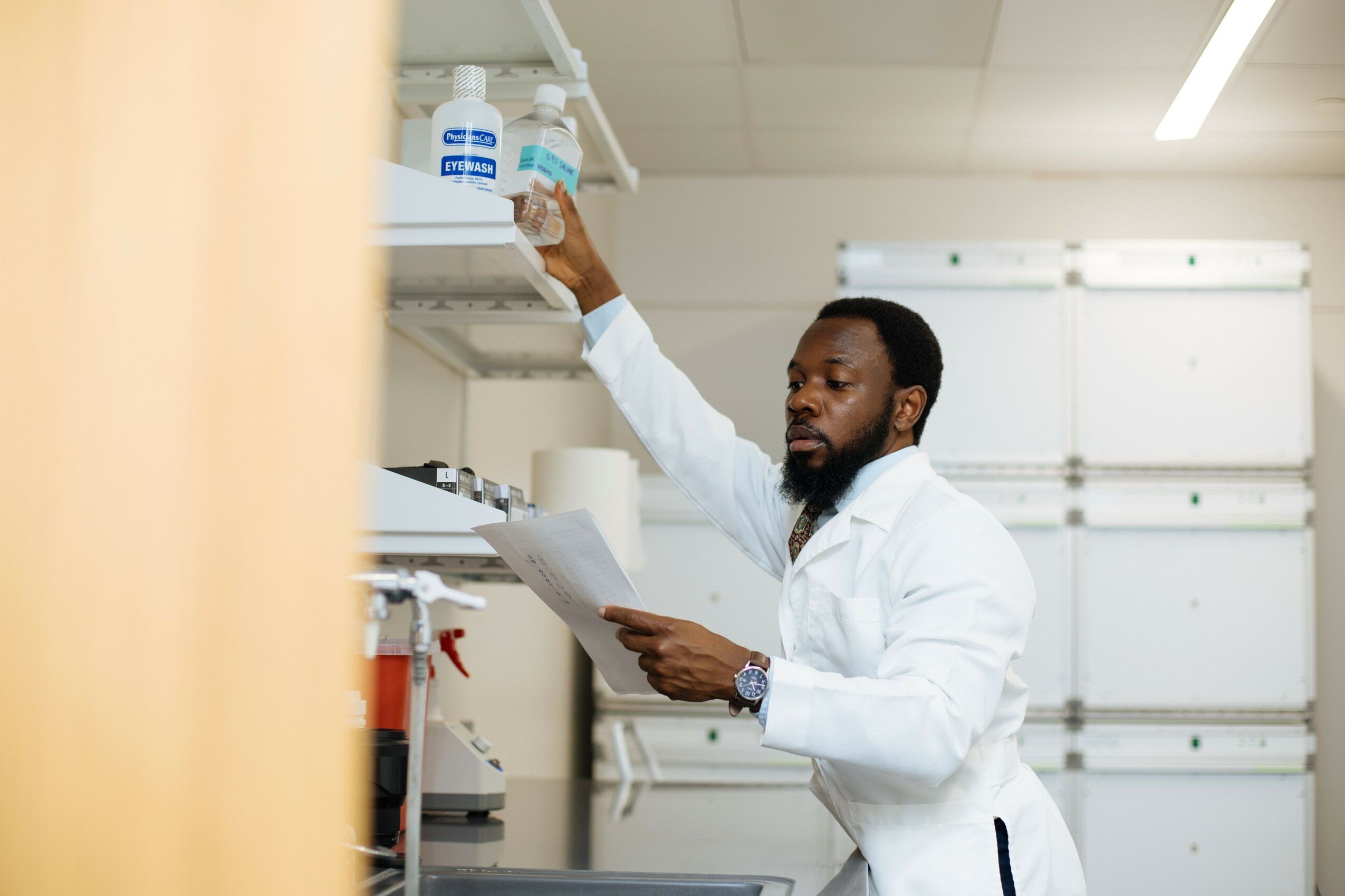 Scientist in a lab coat reaching for bottles on a shelf while holding a clipboard, in a laboratory setting.