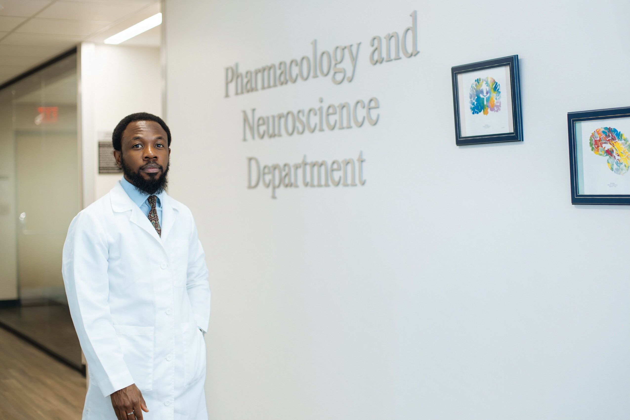 Man in white lab coat standing in front of a wall with text and framed pictures, in a professional setting.