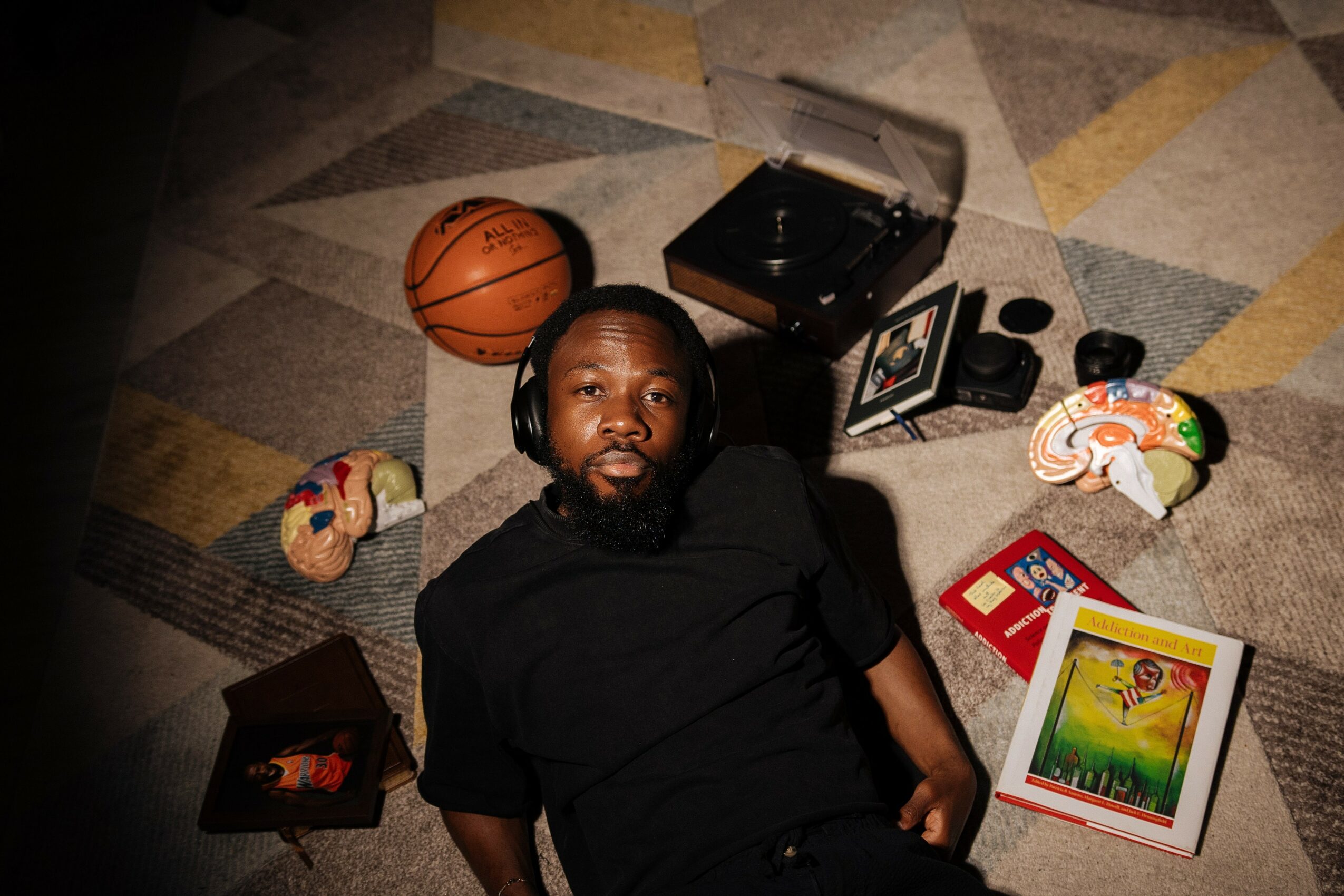 Man with headphones lying on a patterned floor surrounded by a basketball, colorful shoes, a record player, and a book.