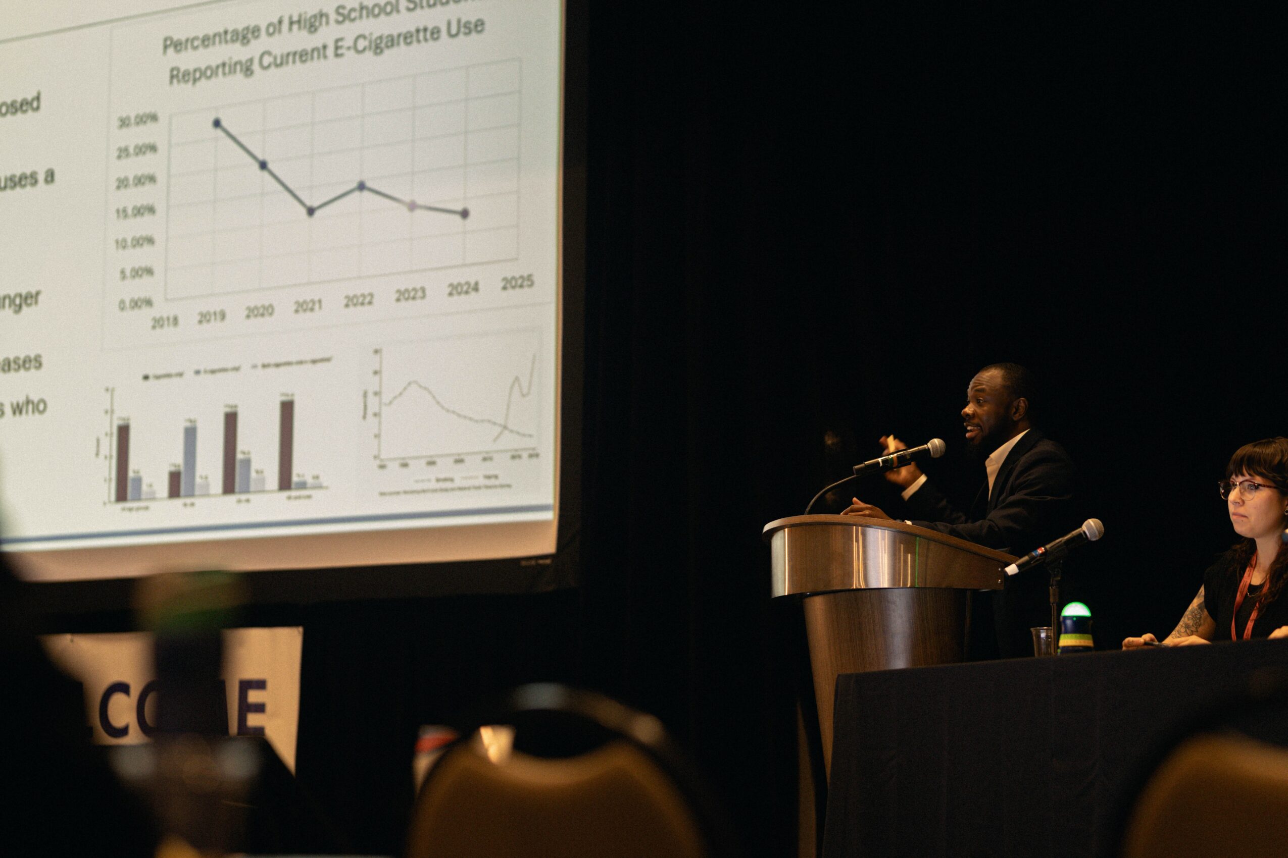 Man speaking at a podium during a presentation with a large screen displaying graphs behind him. A woman is seated nearby.