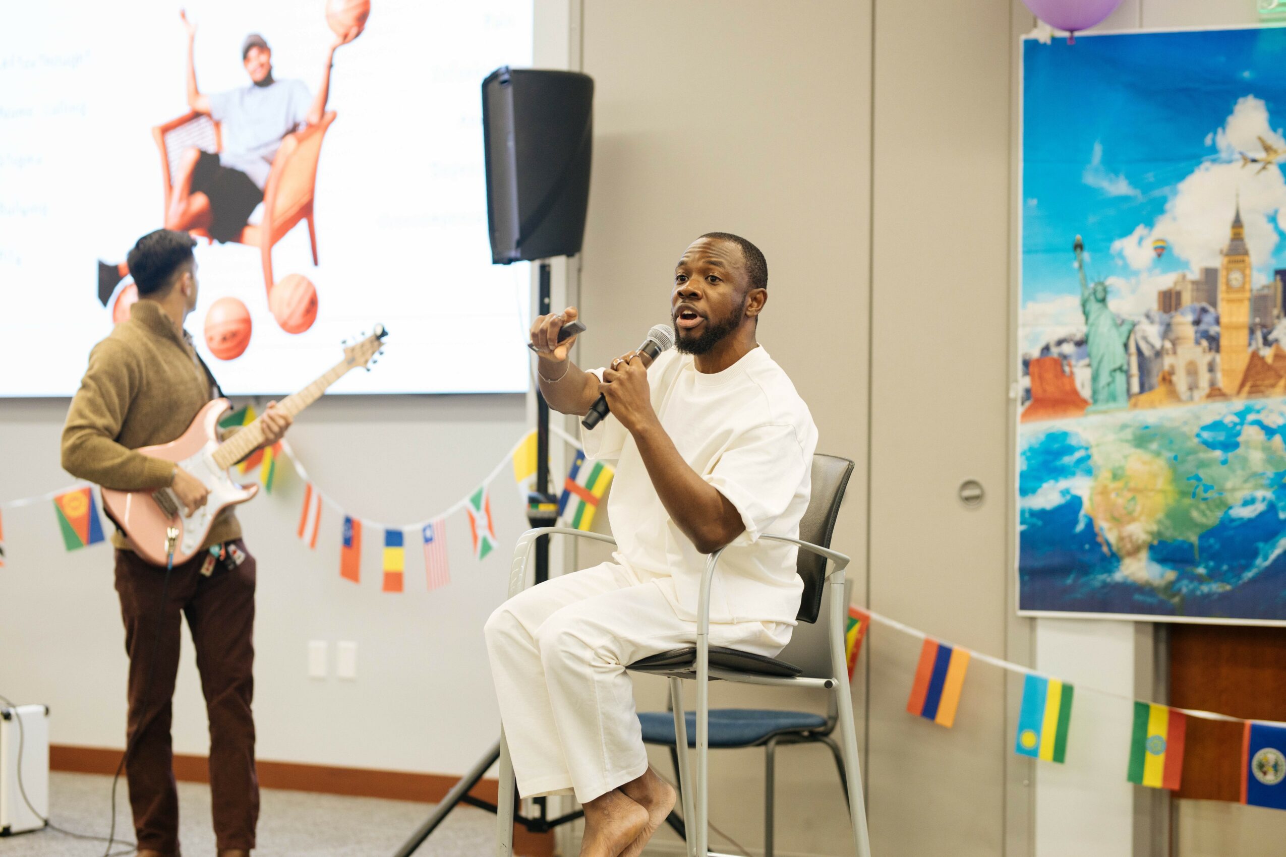 Man singing into microphone while seated, two people playing guitar, colorful decorations, large screen and painting in background.