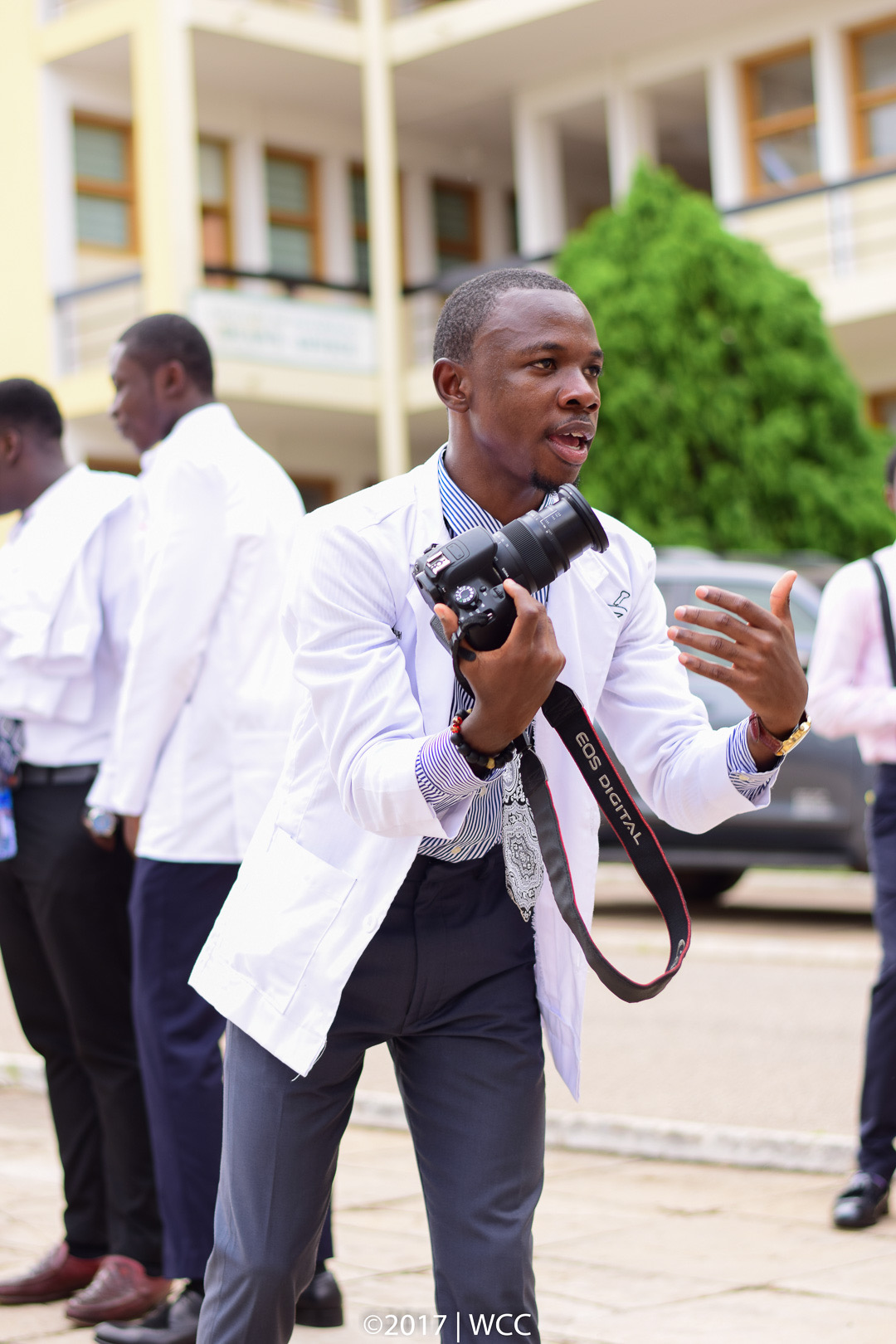 Man holding camera and speaking outdoors with people and a building in background.