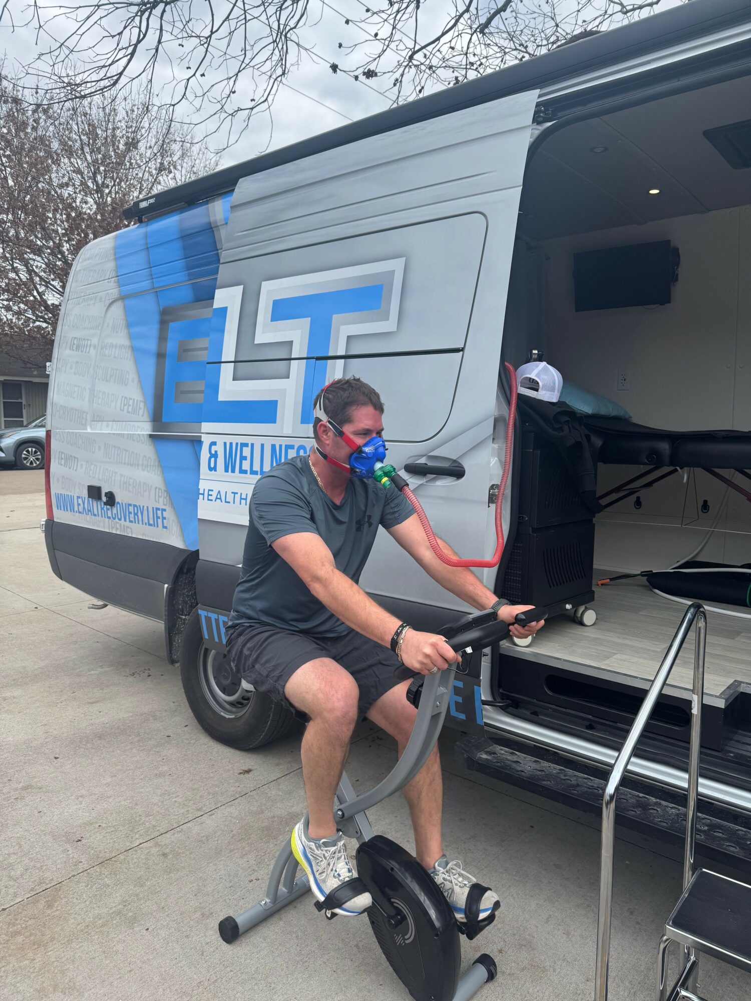 Person wearing a mask and shorts, sitting on a pedal exerciser outside a mobile health clinic, holding a tube connected to a machine.