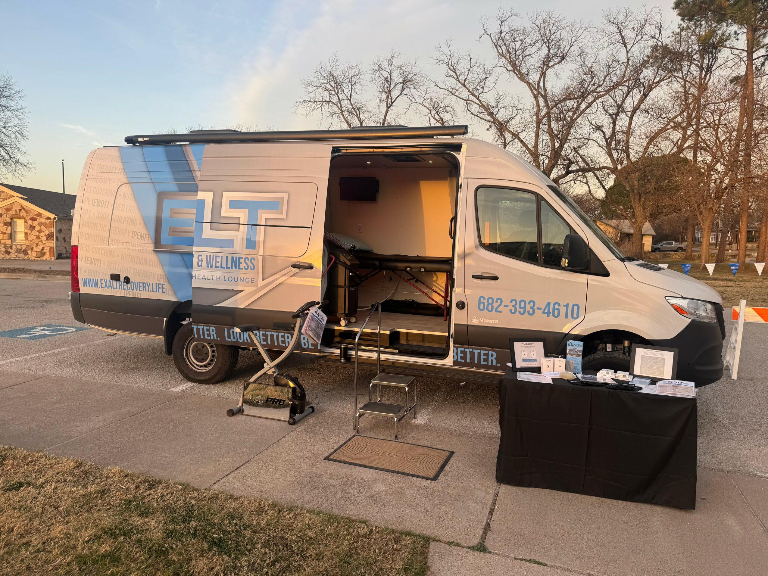 Mobile clinic van with open side door, outdoor setup with chairs, table, and equipment on sidewalk, trees in background.
