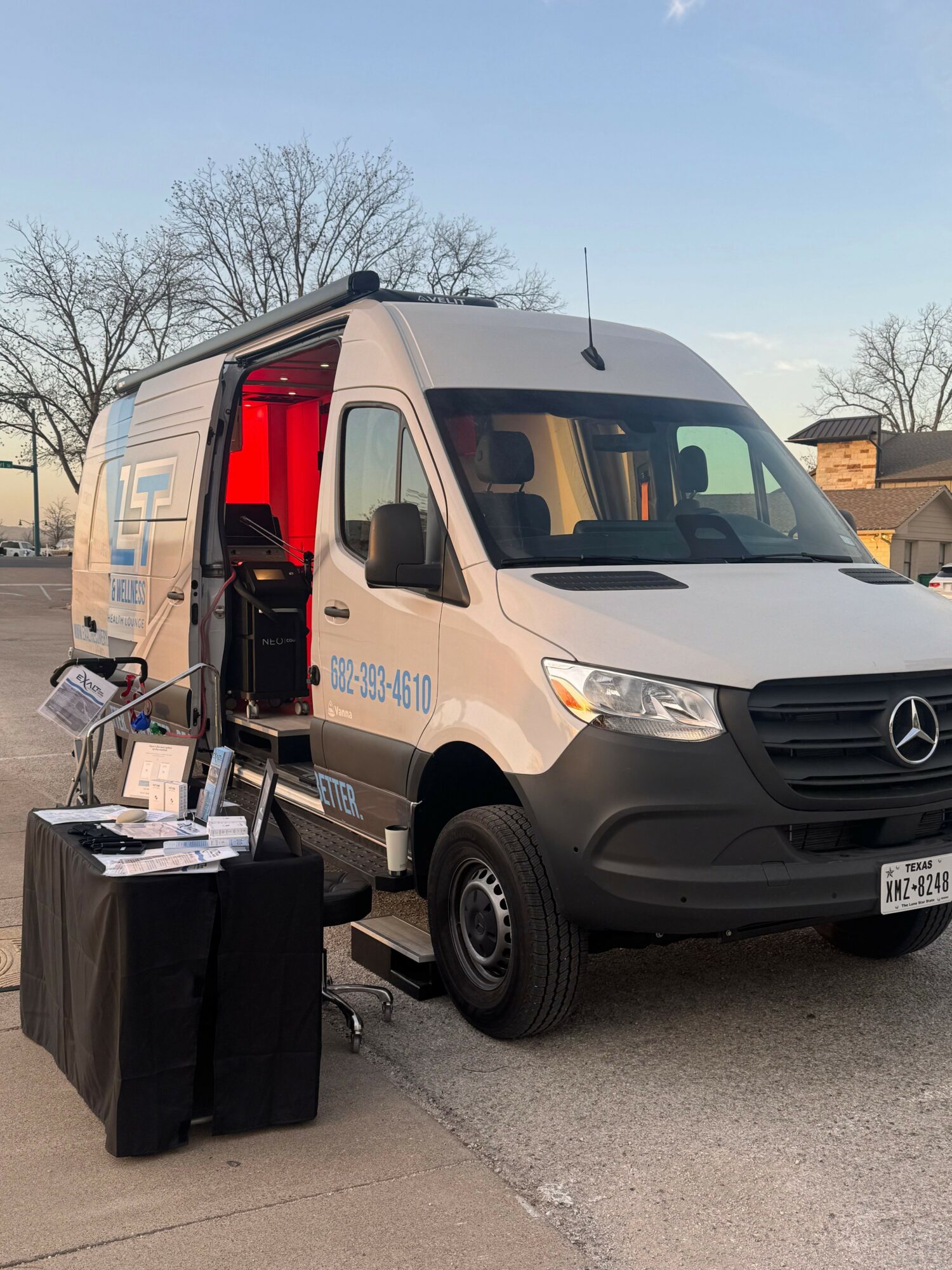 White Mercedes-Benz van parked outdoors with a table and promotional materials beside it, trees and buildings in background.