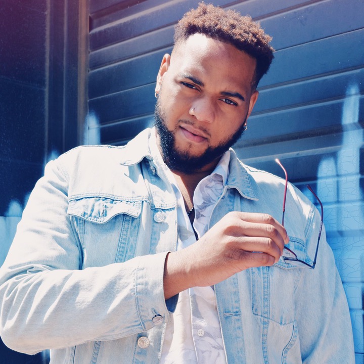 Young man with short hair and beard holding glasses in front of blue wall with graffiti.