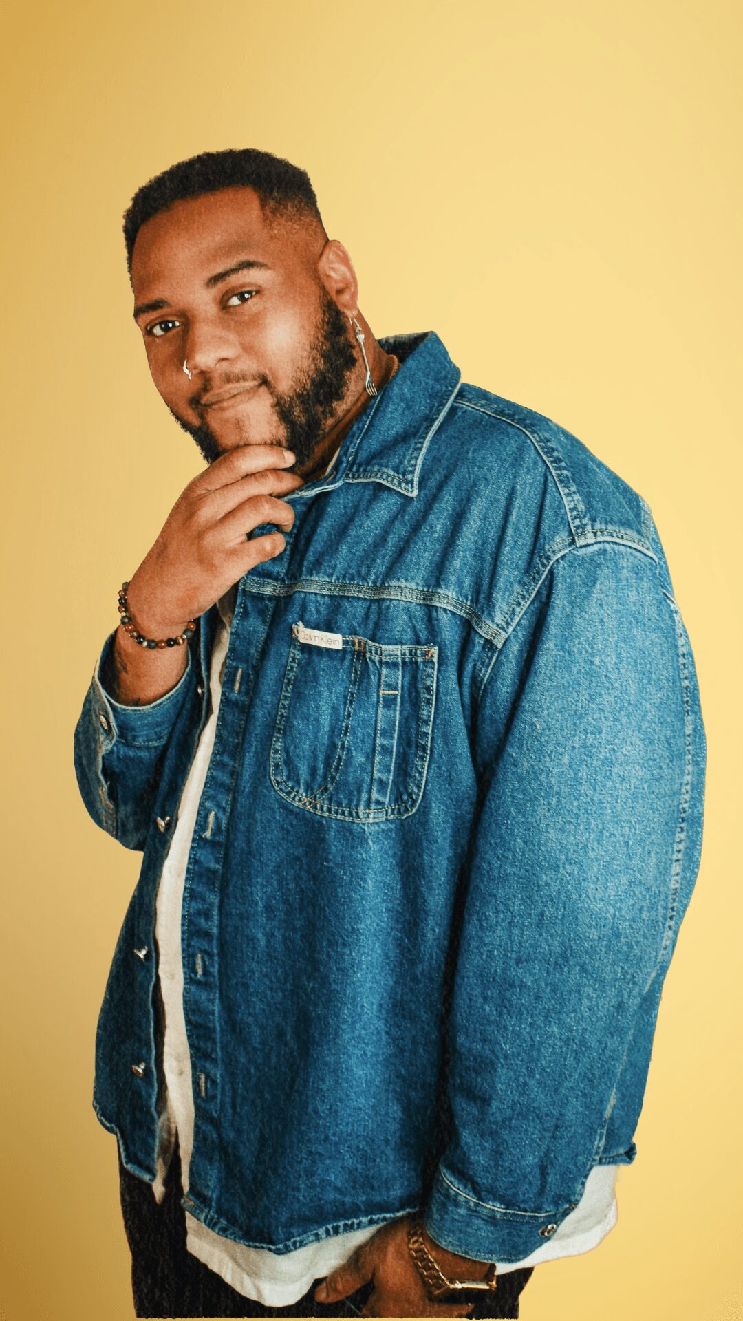 Man with beard wearing denim jacket and white shirt against yellow background.