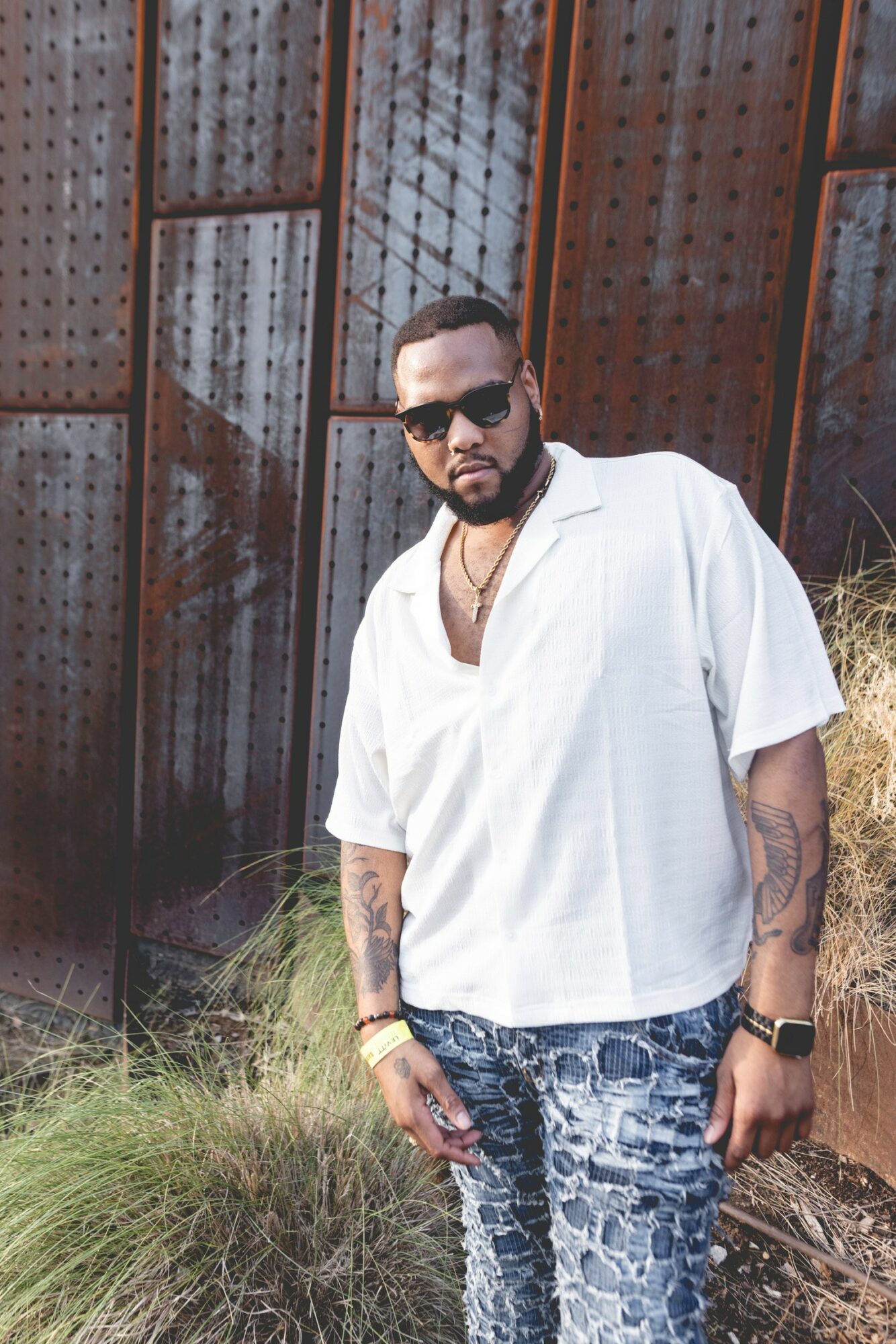 Man with sunglasses, beard, white shirt, patterned jeans, standing outdoors near a metal fence.