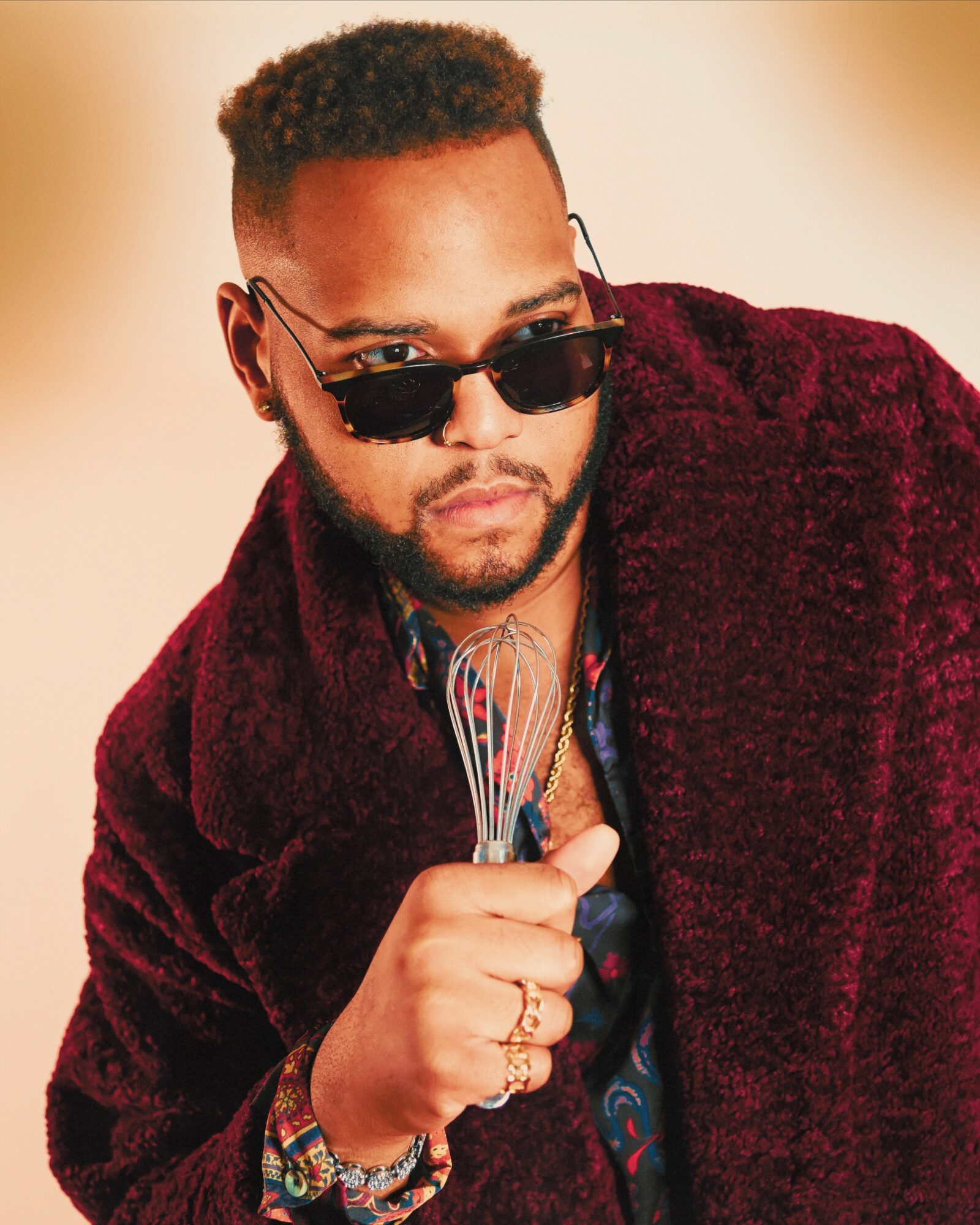 Man with sunglasses and a maroon coat holding a whisk, looking to the side.