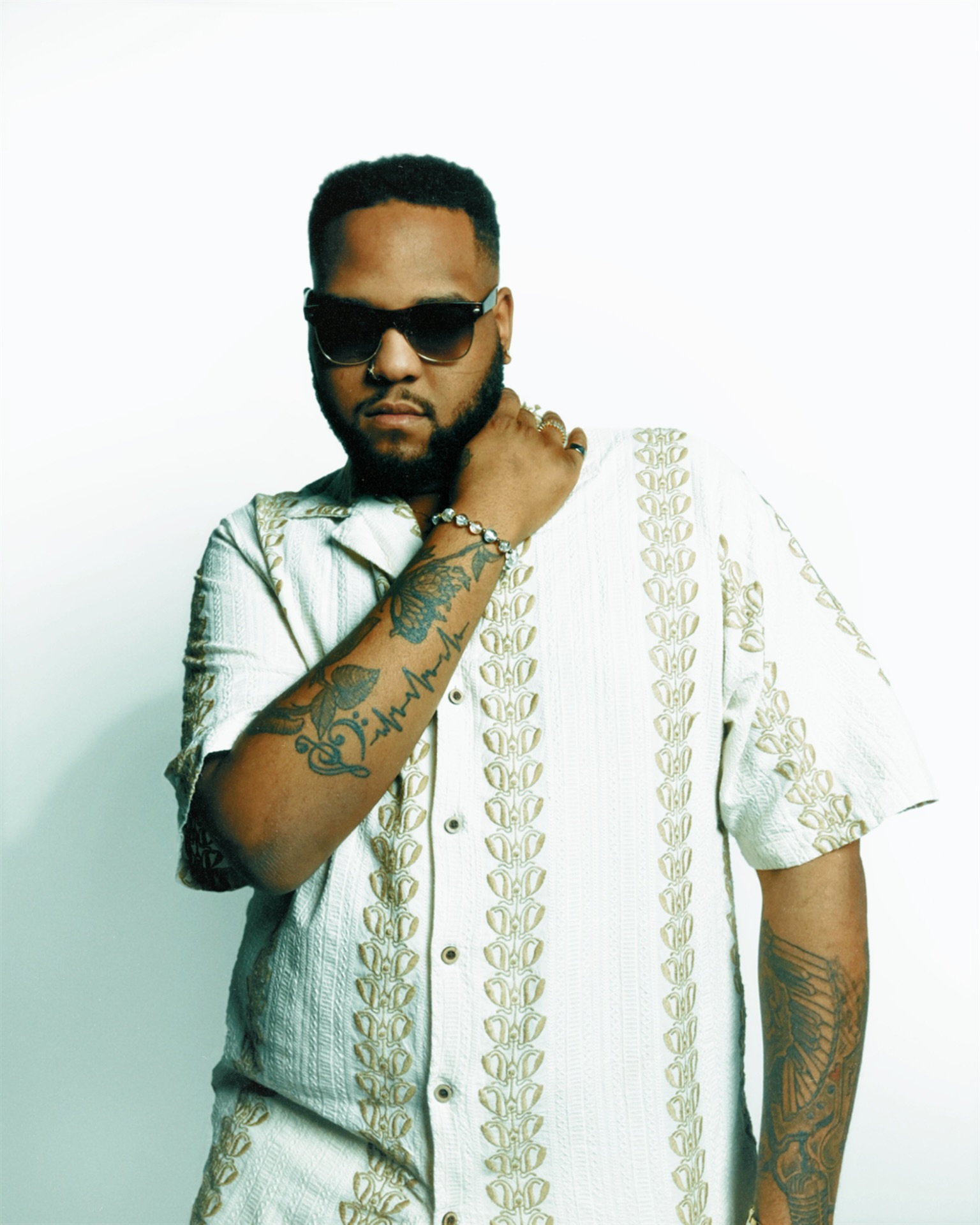 Man with sunglasses and tattoos wearing a patterned shirt, posing with hand near face against a plain background.