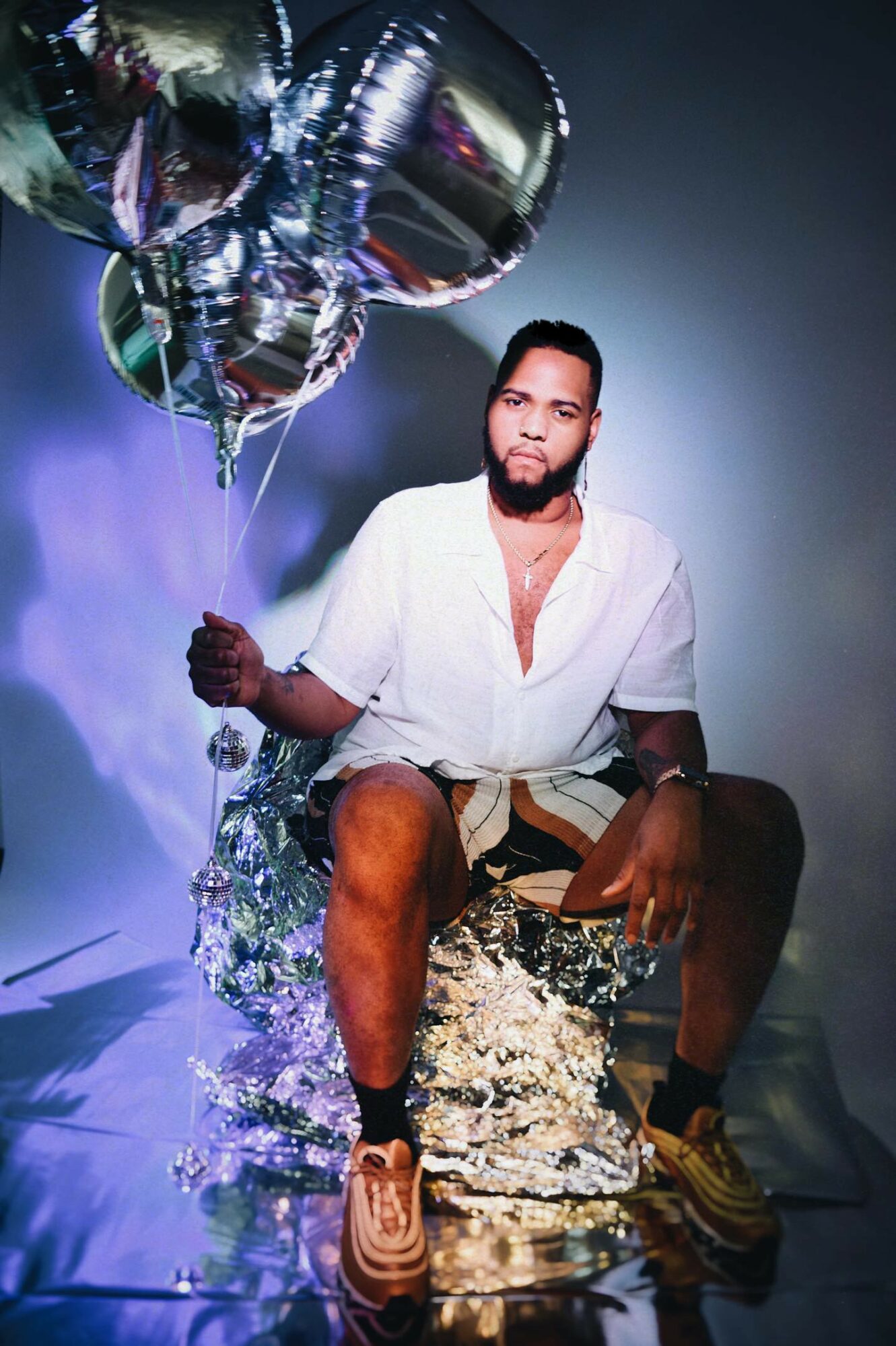 Man sitting on a reflective surface with balloons, water splashing around, wearing a white shirt and shorts.