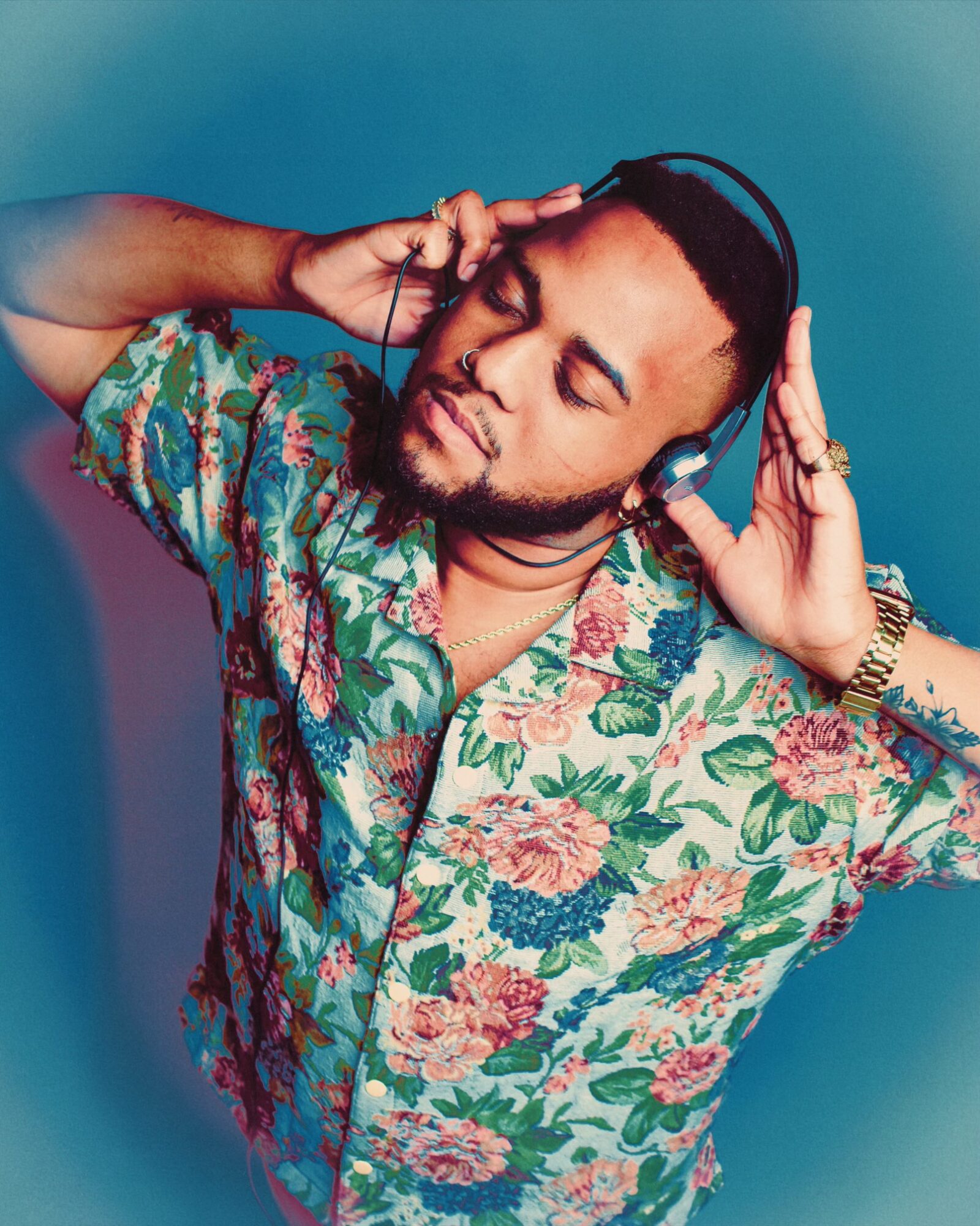 Man with closed eyes wearing headphones and a colorful floral shirt, holding headphones with both hands.