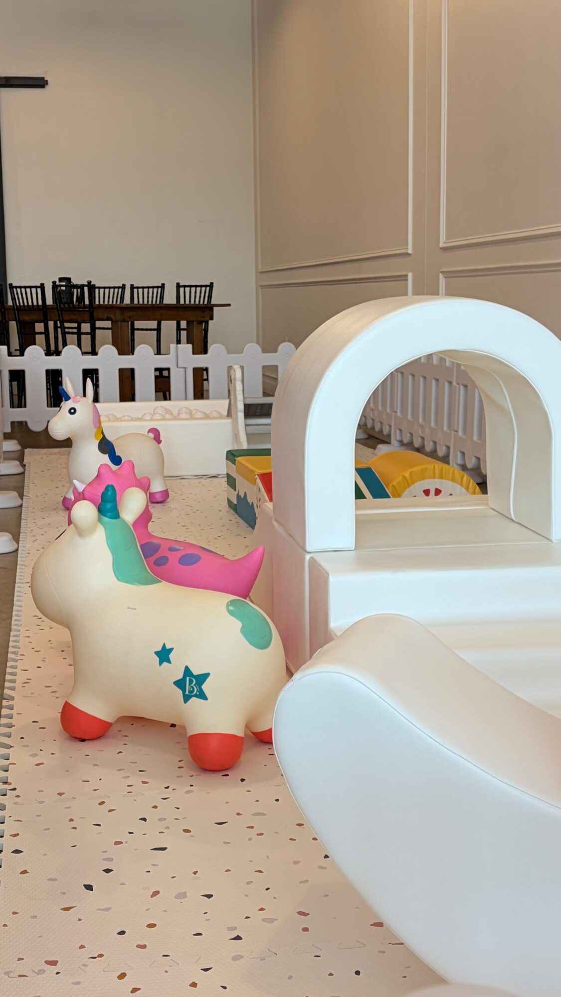 Children's play area with unicorn-shaped ride-on toy, white arch structure, and colorful toys, in a room with white walls and furniture.