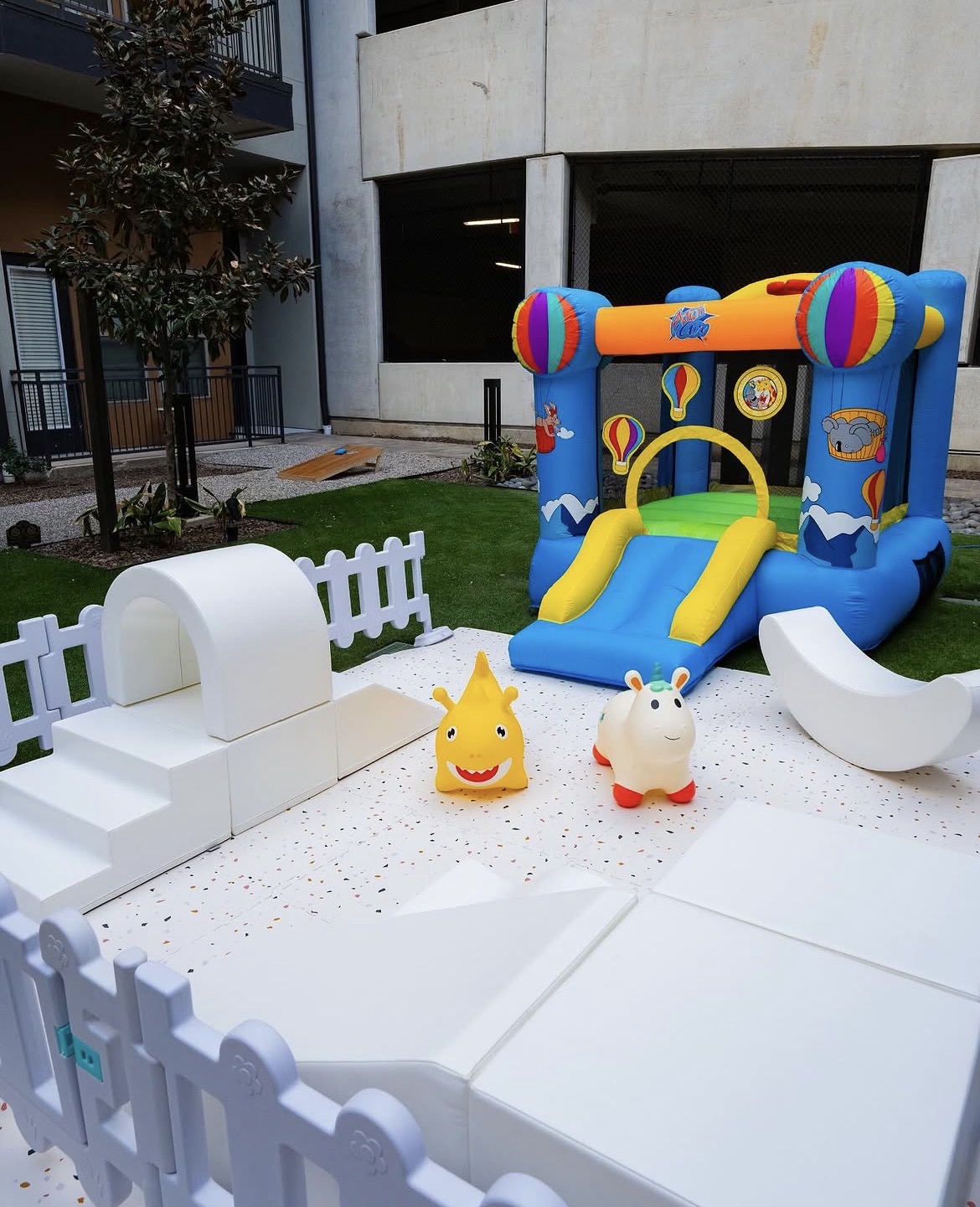 Colorful inflatable bounce house with slide, two plush toys, and a white fence in an outdoor area.