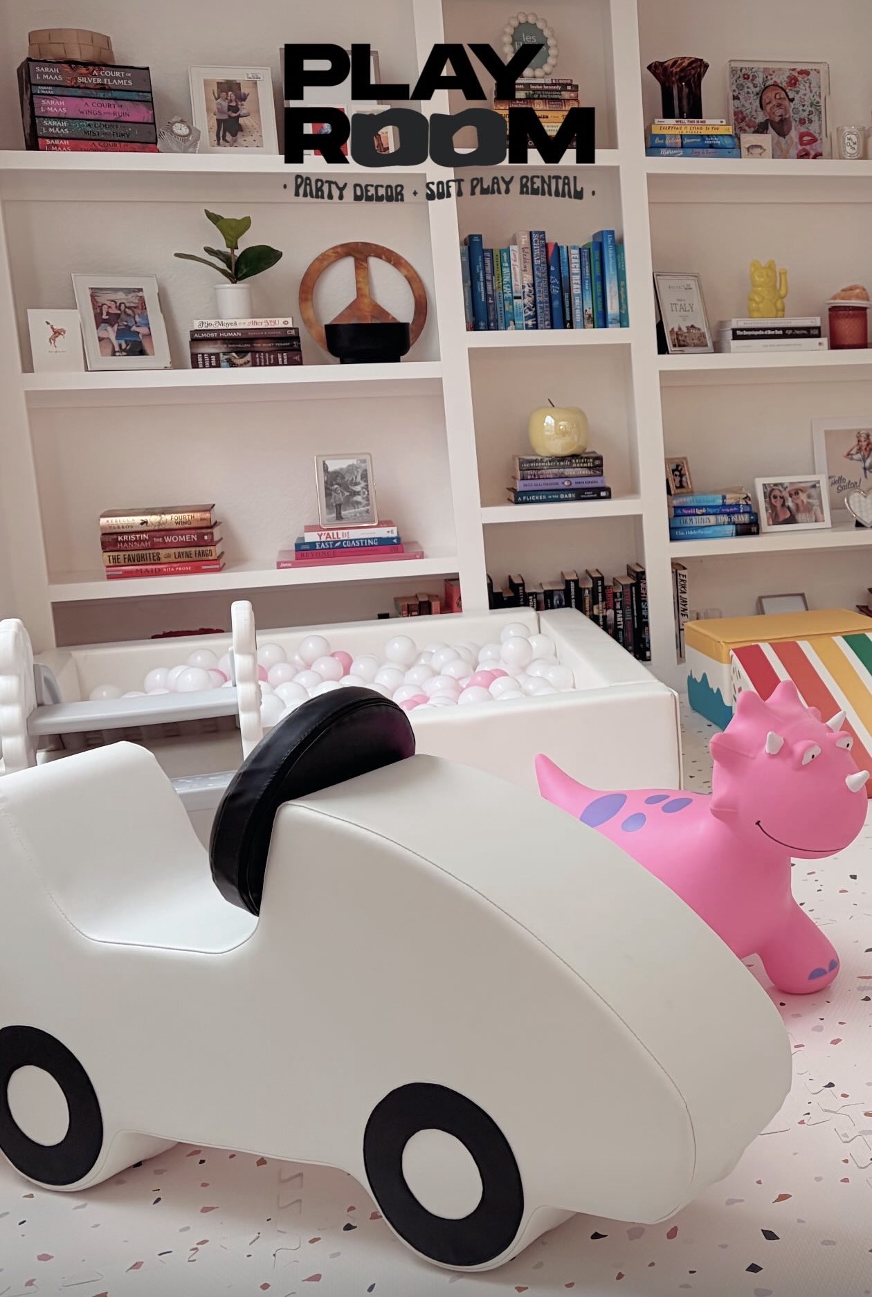 Children's playroom with white car-shaped toy, pink inflatable horse, and white bookshelf with books and decorations.