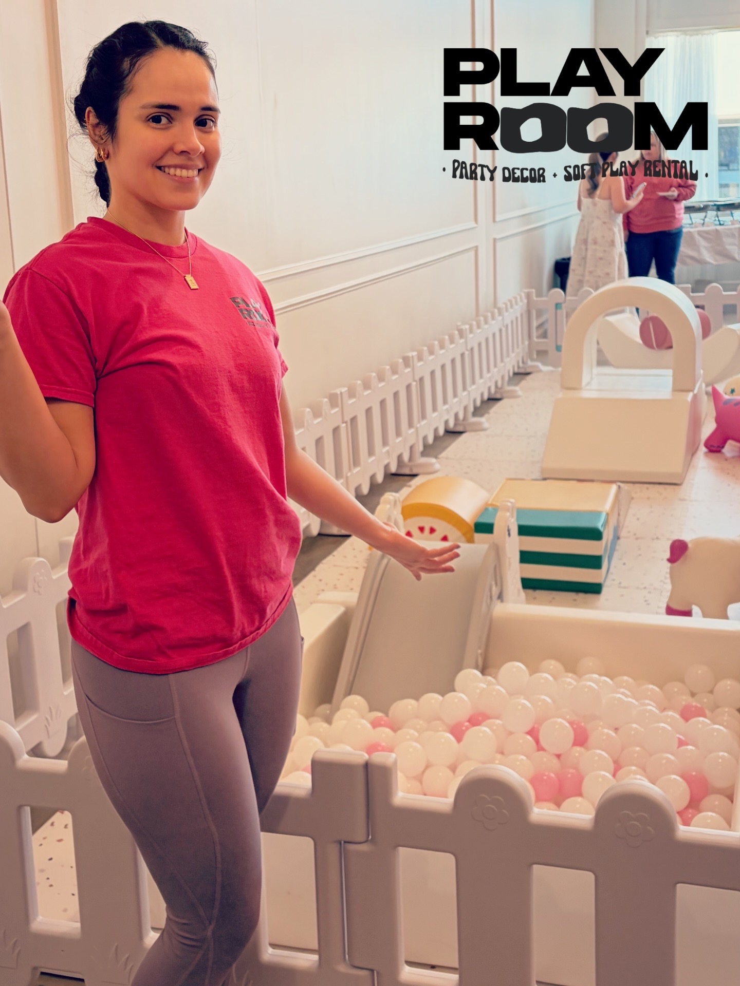 Woman in pink shirt standing next to a ball pit with white fence, smiling indoors.
