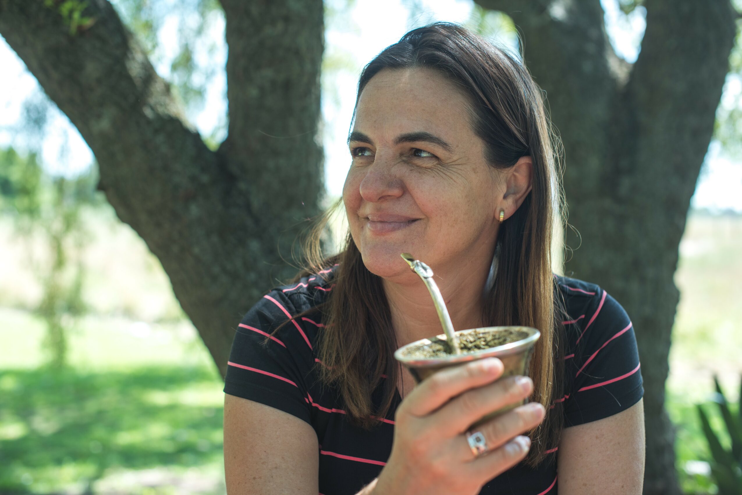 Woman with dark hair smiling outdoors holding a cup with a straw, trees in background.