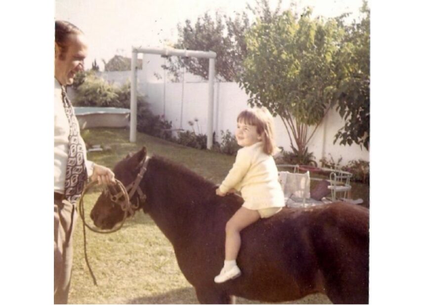 Child sitting on a horse with an adult holding the reins outdoors in a garden.