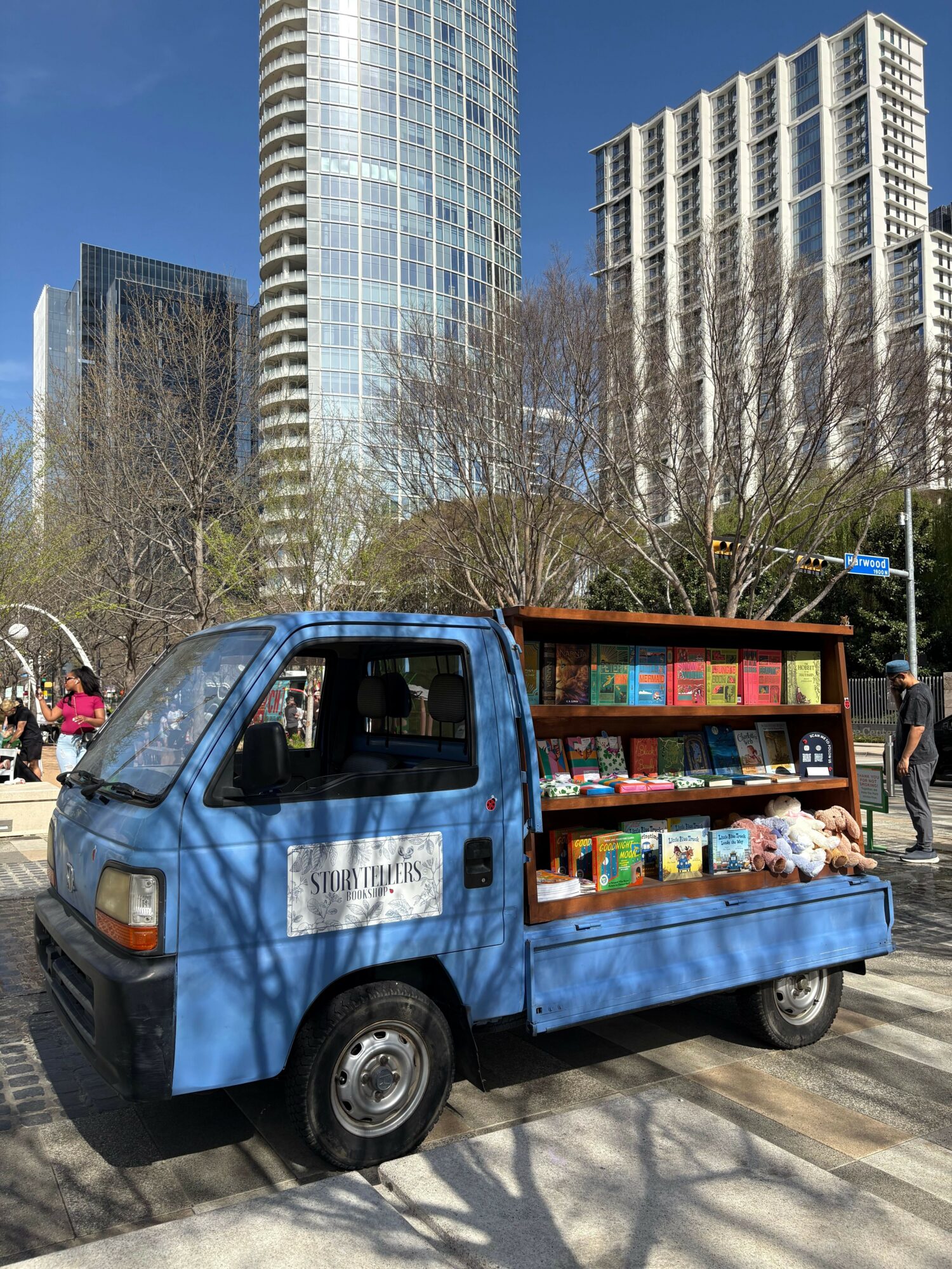 Small blue mobile bookstore truck parked outdoors with tall modern buildings in the background.
