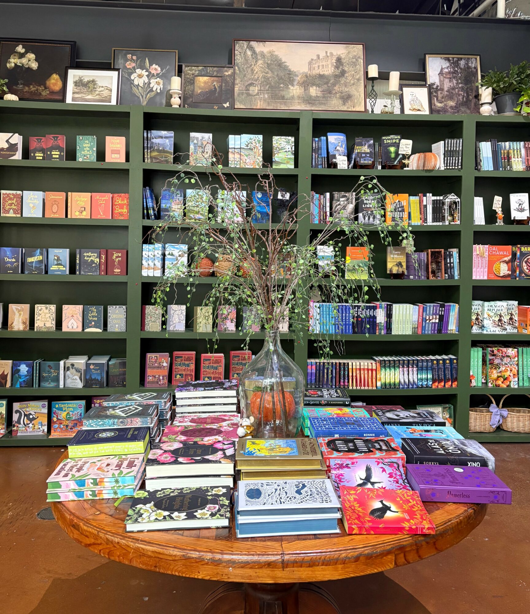 Round wooden table with books and a glass vase holding a plant, in front of a bookshelf filled with books and framed pictures.
