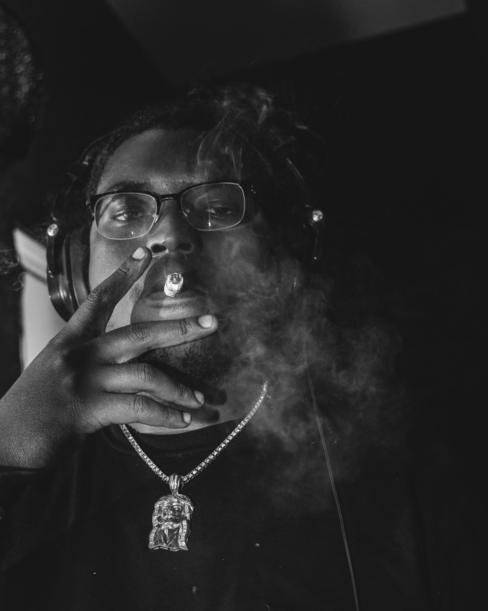 Person wearing glasses and headphones, smoking with smoke visible, holding fingers near face, wearing a chain necklace.