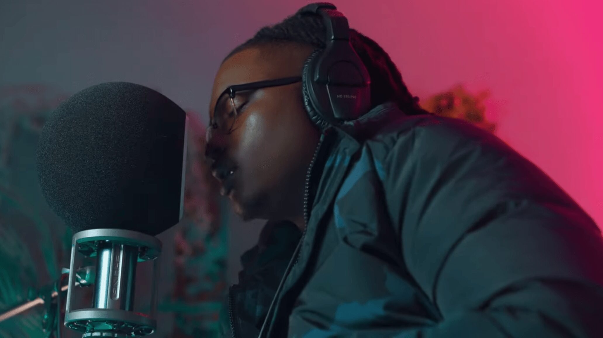 Person wearing headphones and glasses, speaking into a microphone in a recording studio with colorful lighting.