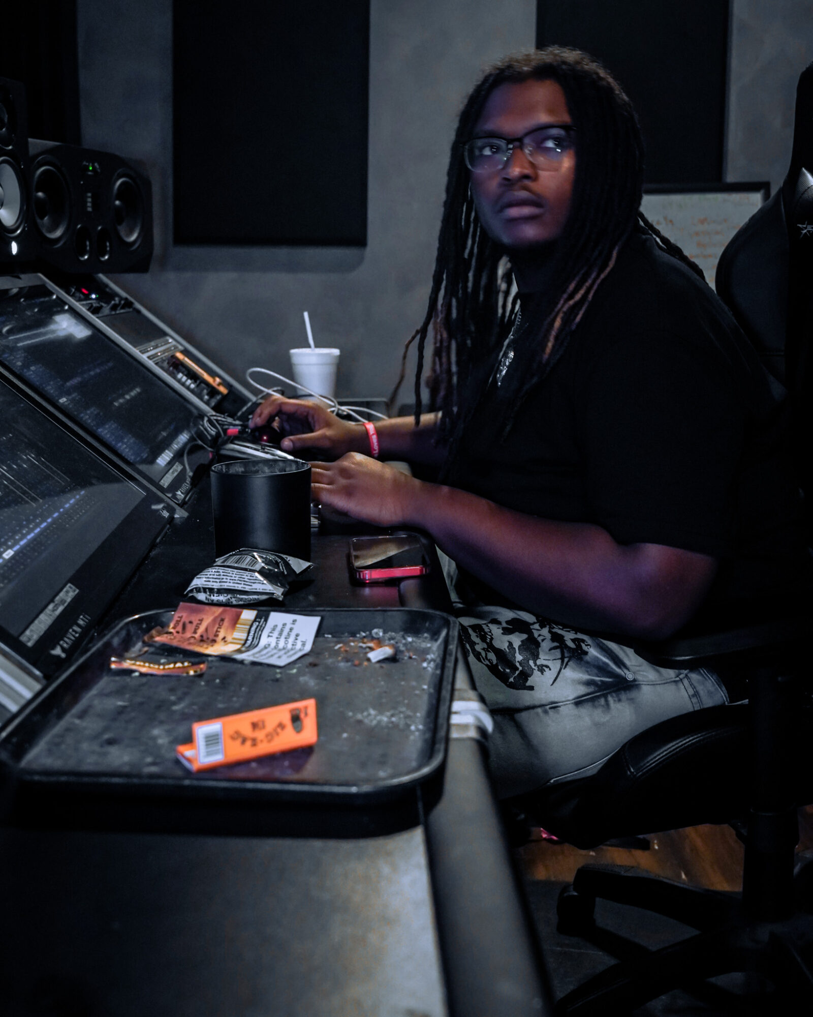 Person with glasses and long hair sitting at mixing console in recording studio, with drink and snacks nearby.