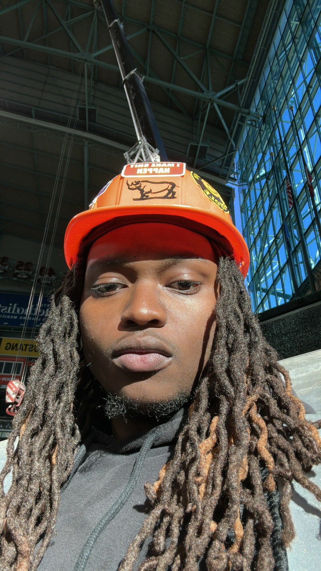 Person with long dreadlocks wearing an orange construction helmet and red cap inside a building with large windows.