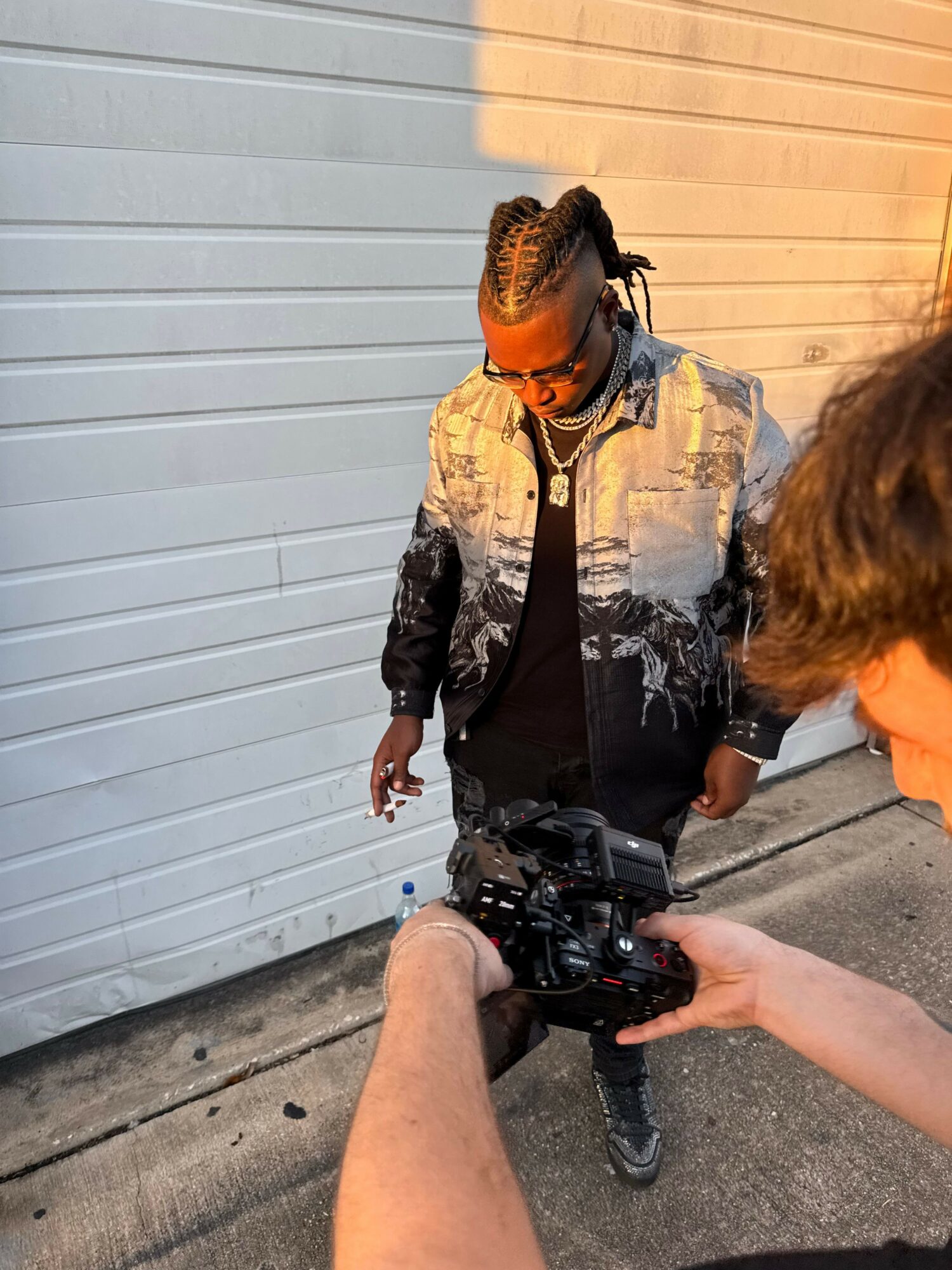 Person with braided hair and sunglasses looking down, standing outside near a white garage door, with a camera in foreground.