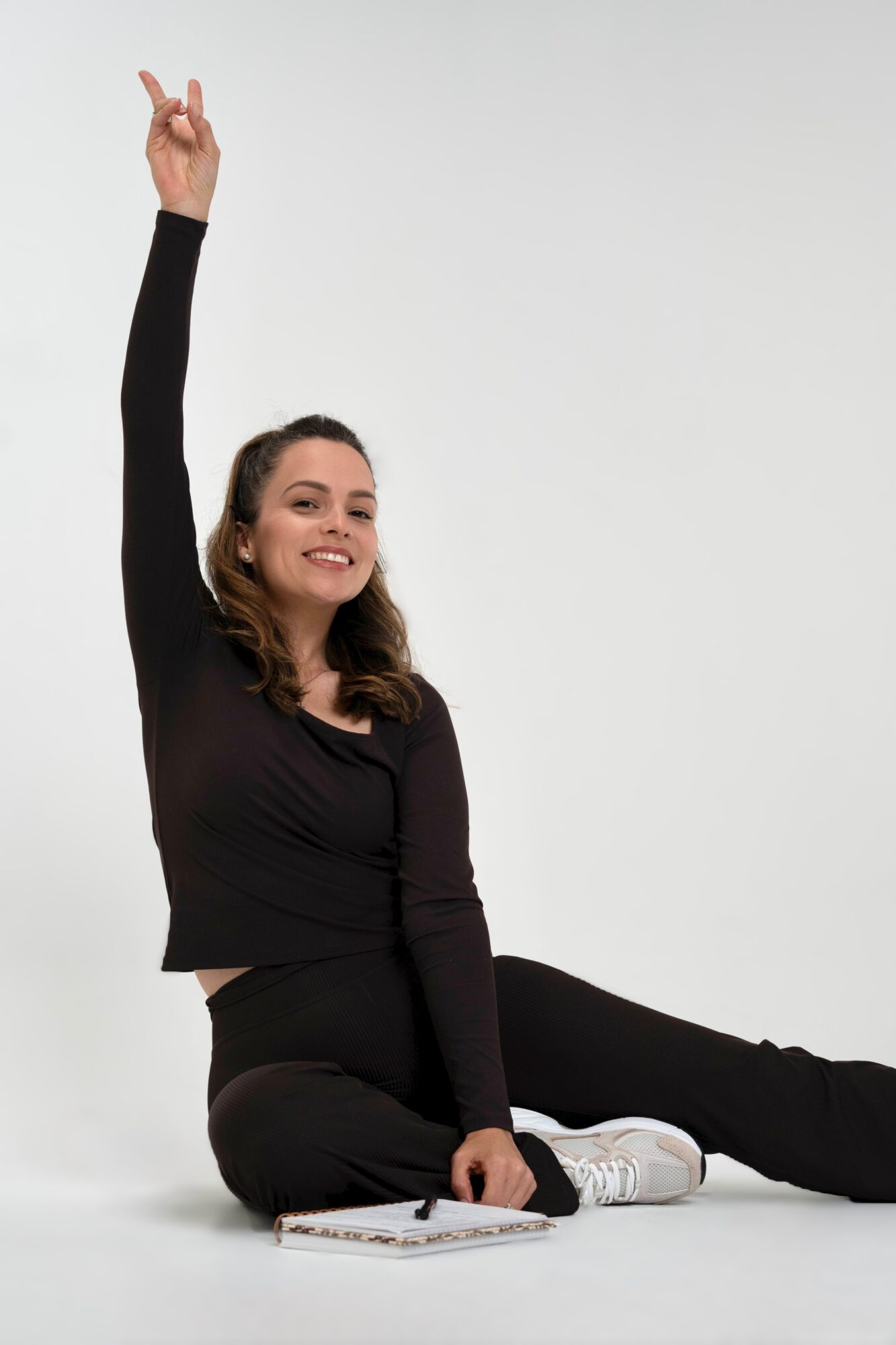 Young woman sitting on the floor with one arm raised, smiling, against a plain background.