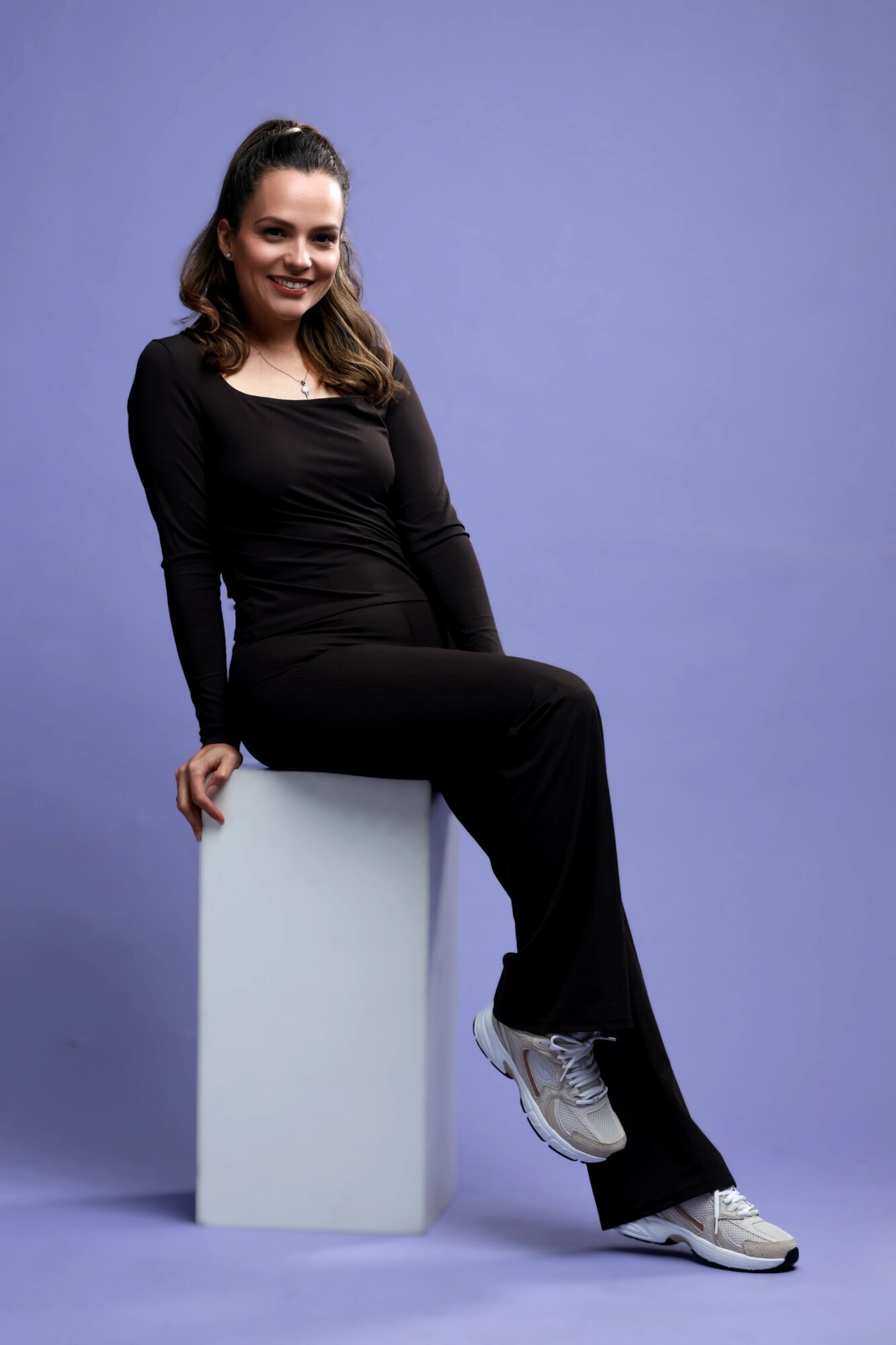 Young woman in black outfit sitting on a white block against a purple background, smiling.