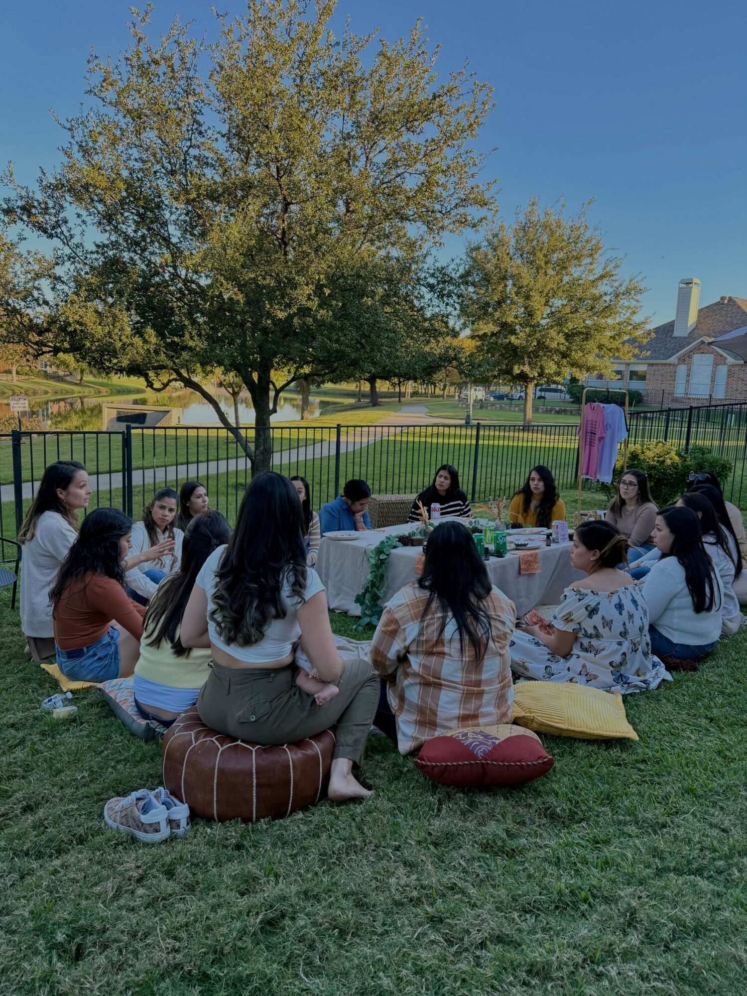 Group of people sitting outdoors on grass around a table with food and drinks, trees and a house in background.
