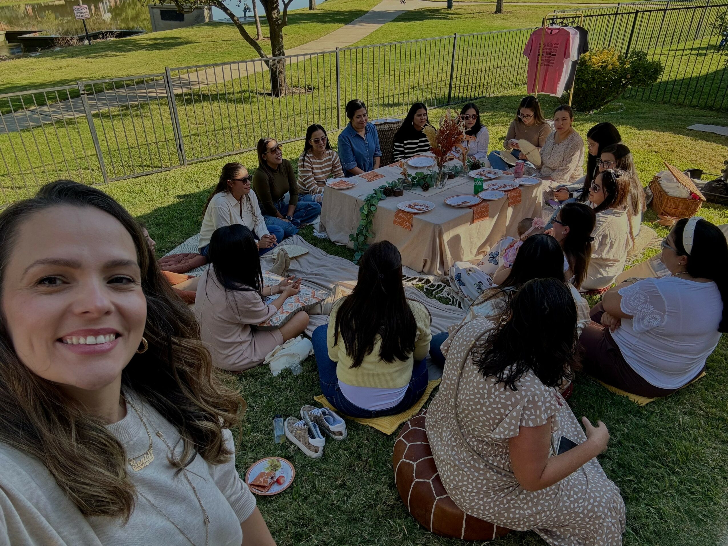 Group of women and children sitting around a table outdoors, enjoying a meal in a park-like setting.