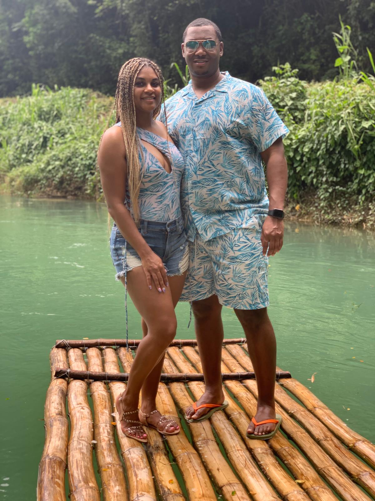 A man and woman stand on a wooden raft by a river, surrounded by greenery, wearing summer clothes.