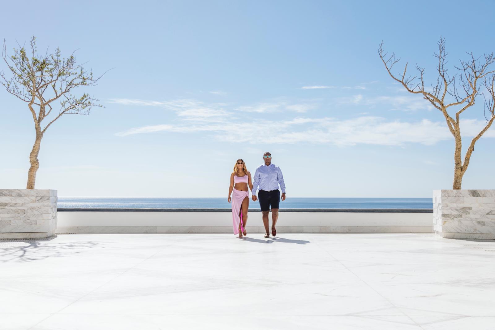 Two people walk along a promenade with trees in large planters, overlooking the ocean under a blue sky.