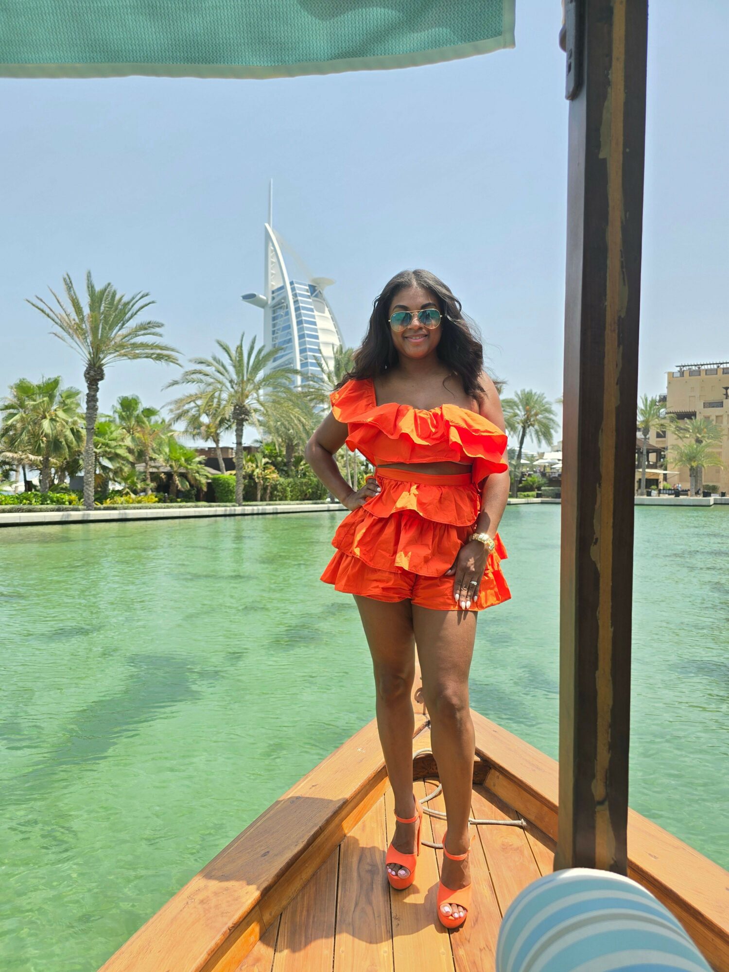 Woman in orange dress standing on boat with water, palm trees, and a building in background.