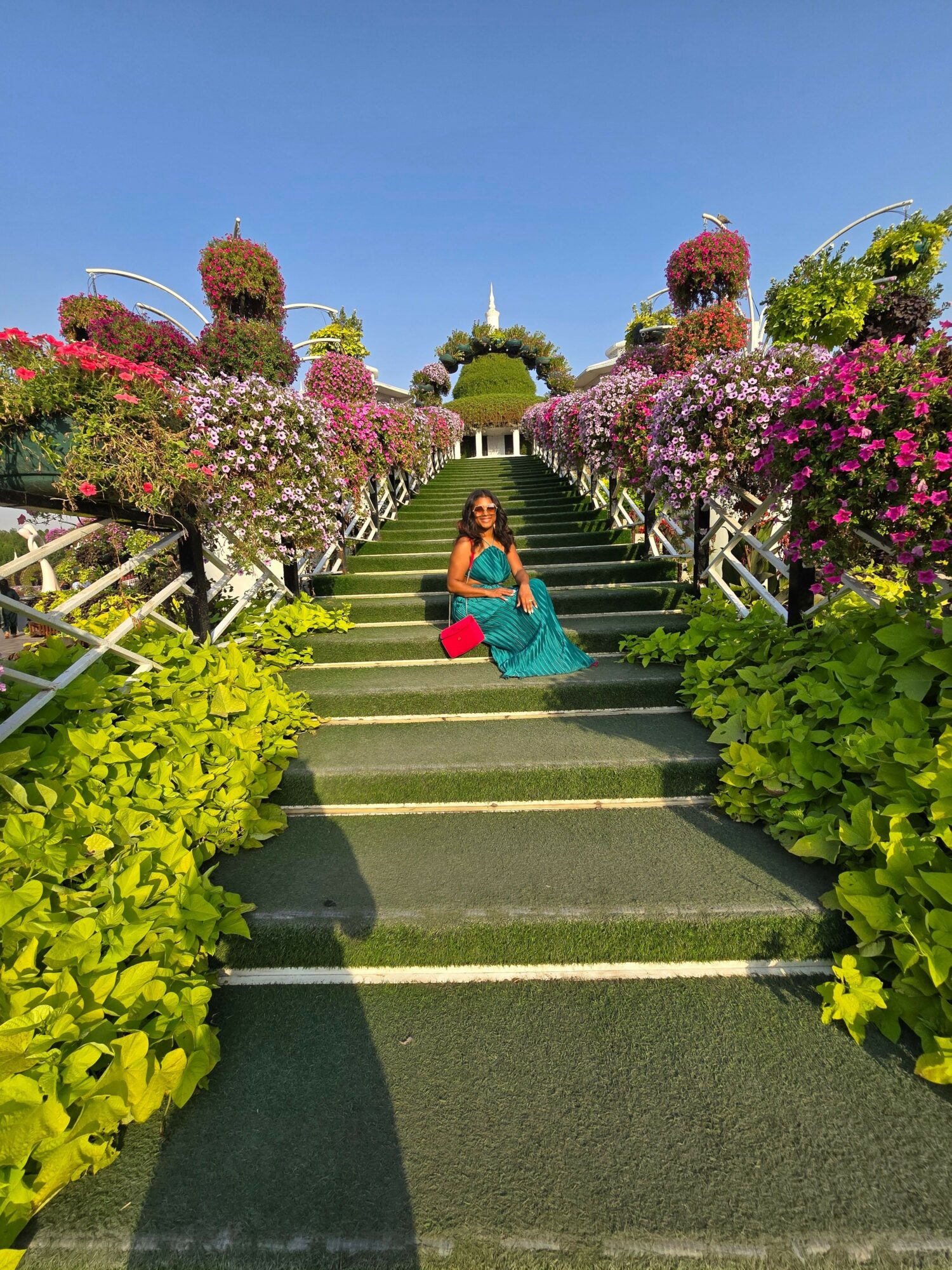 Woman in blue dress sitting on steps surrounded by colorful flowers and greenery, clear blue sky above.